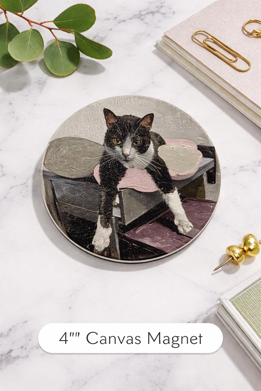 Custom Pet Portrait Magnet with a textured brushstroke portrait of a black and white cat, styled flat lay on a white marble surface beside a book, paper clips and a green leaf sprig