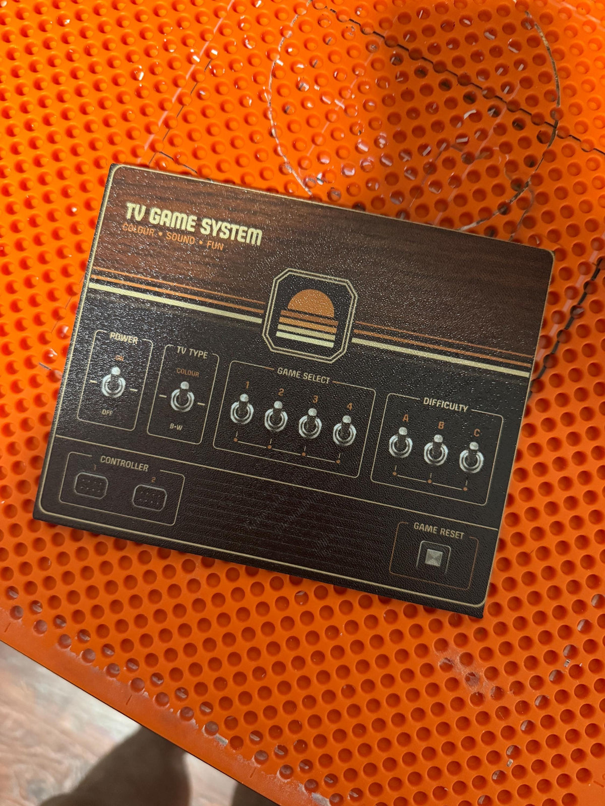 Retro Woodgrain Steam Machine Faceplate – 70s Atari Inspired Console Cover, flat lay of the uninstalled woodgrain faceplate panel on an orange perforated plastic surface