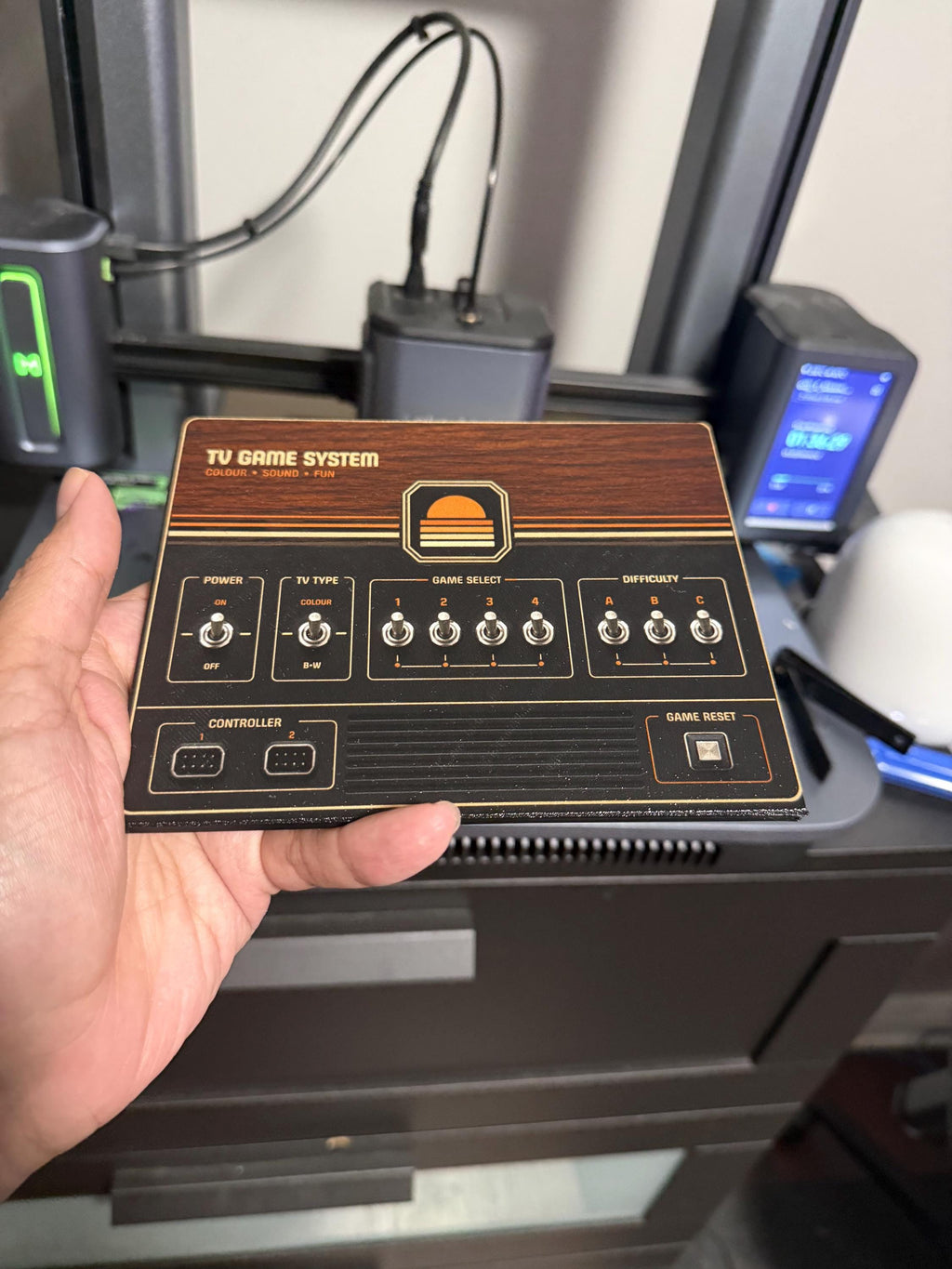 Retro Woodgrain Steam Machine Faceplate – 70s Atari Inspired Console Cover, hand holding the uninstalled woodgrain faceplate panel in front of a 3D printer on a workshop desk