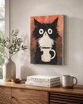 Funny Tuxedo Cat Coffee Canvas Art print of a wide-eyed tuxedo cat holding a coffee mug, hanging on a wall above a wooden console table styled with stacked books, a ceramic vase and a coffee mug