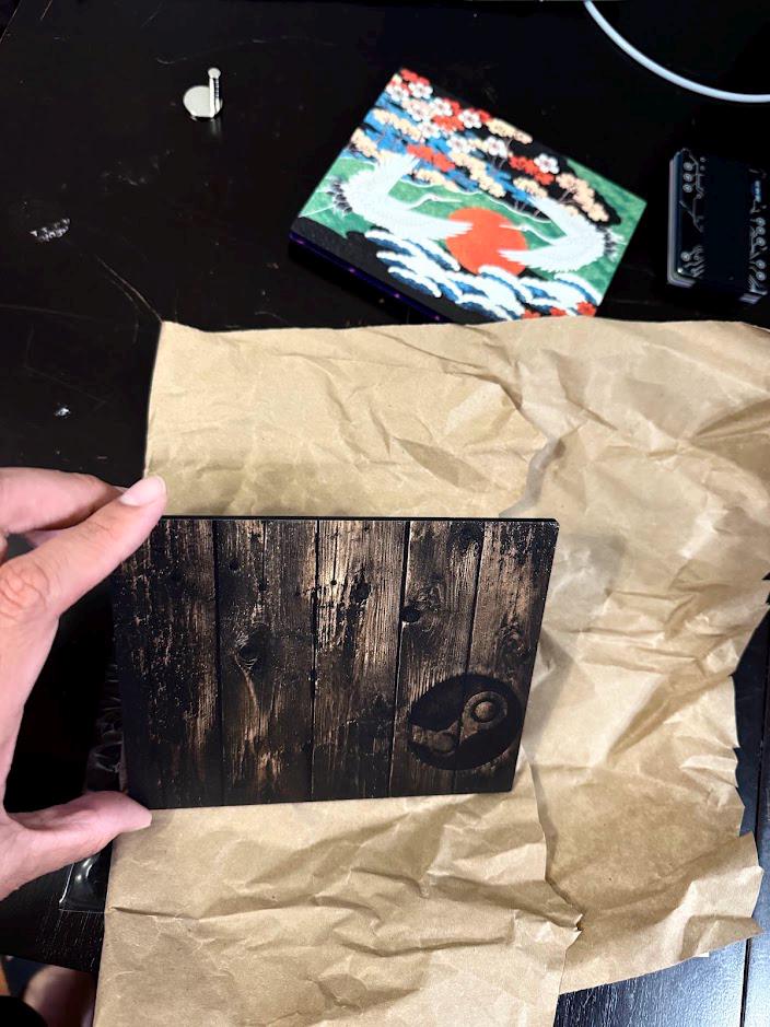 Custom Steam Machine Faceplate, Build your own Personalized Magnetic Cover Plate, Your Photo or Art UV Printed, Gamer Gift Valve Steam, sample printed faceplate panel with a dark wood plank pattern and small circular logo held in a hand over brown packaging paper, with other sample panels on the desk behind