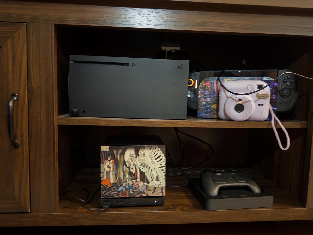 Steam Machine Faceplate, Kuniyoshi Ukiyo-e Skeleton Art, UV Printed Panel with the giant skeleton ukiyo-e artwork, shown installed on the console on the lower shelf of a wooden media cabinet with a game controller on a charging dock and a console and instant camera above