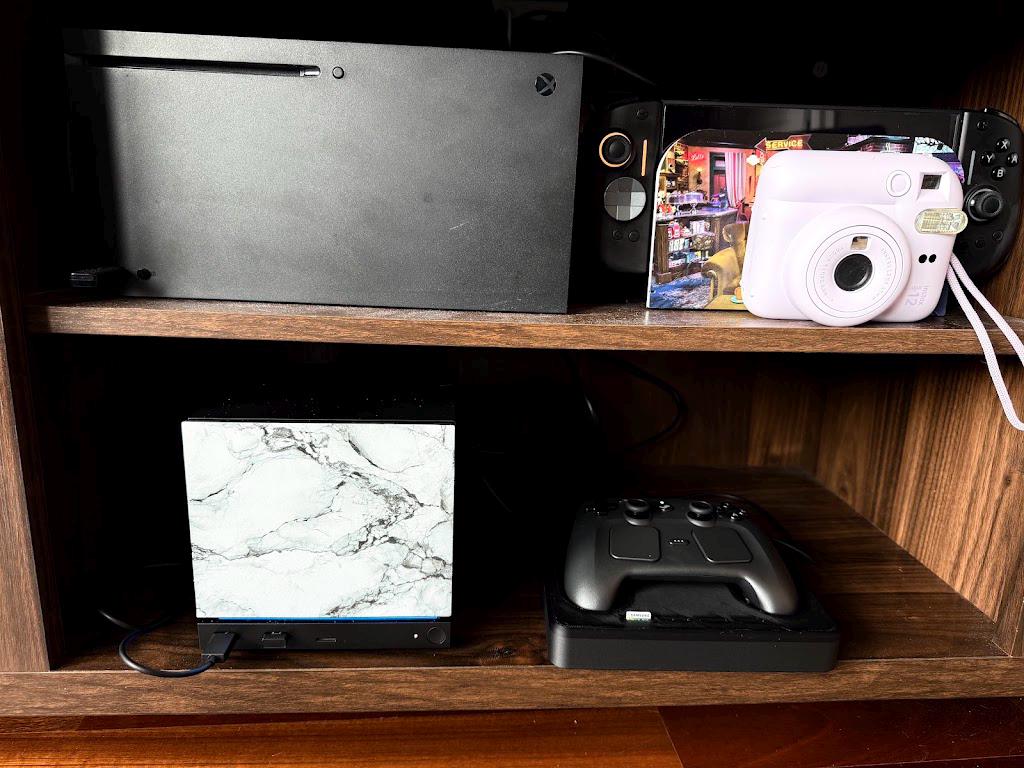 White Marble Steam Machine Faceplate | Magnetic UV Printed Panel, Minimal Desk Decor, wider view of the Steam Machine with the white marble faceplate installed on a wooden shelf next to a Steam controller on its charging dock, with an Xbox console and an instant camera on the shelf above