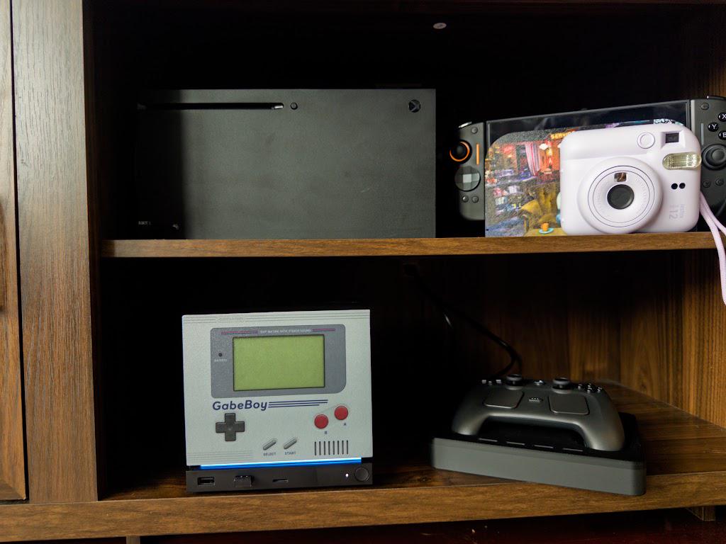 GabeBoy Retro Handheld Style Steam Machine Faceplate on the console, wide shot of a wooden entertainment shelf with a game controller on a stand and other consoles above