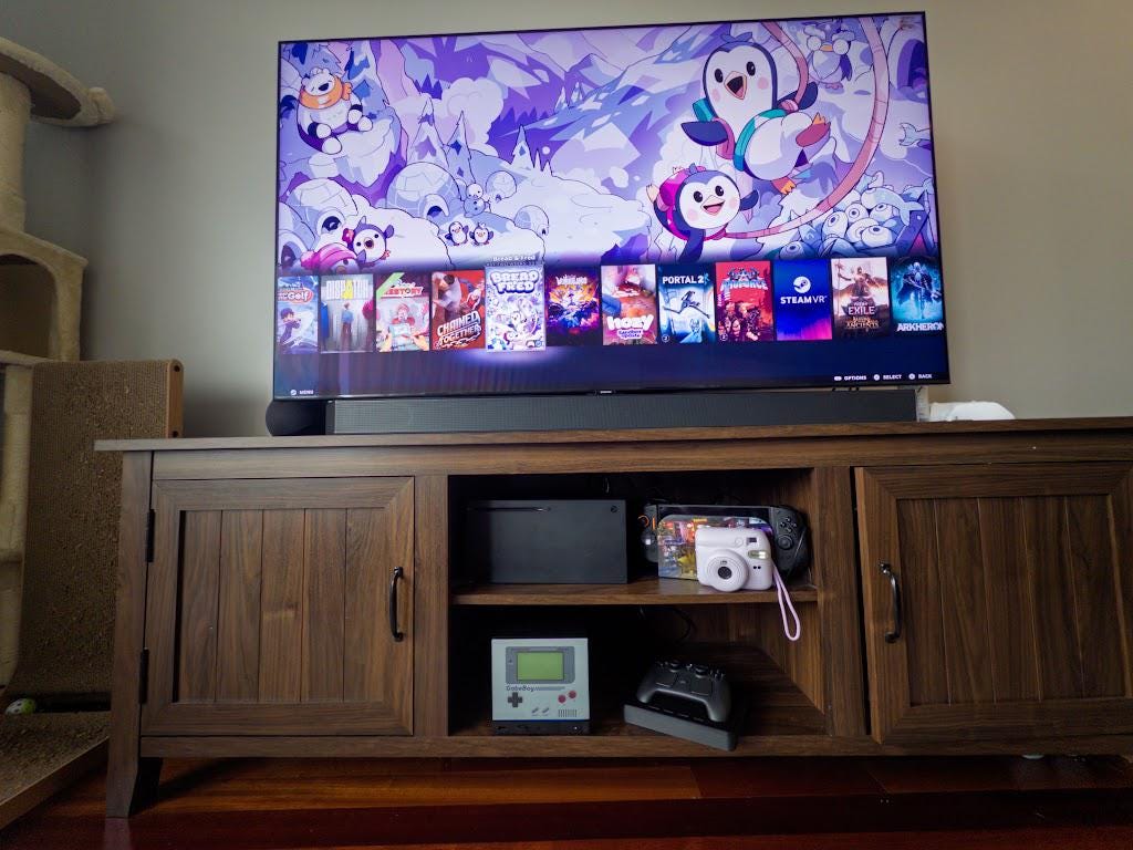 GabeBoy Retro Handheld Style Steam Machine Faceplate on the console, wide living room shot of the wooden entertainment stand with the console visible on the middle shelf beneath a TV