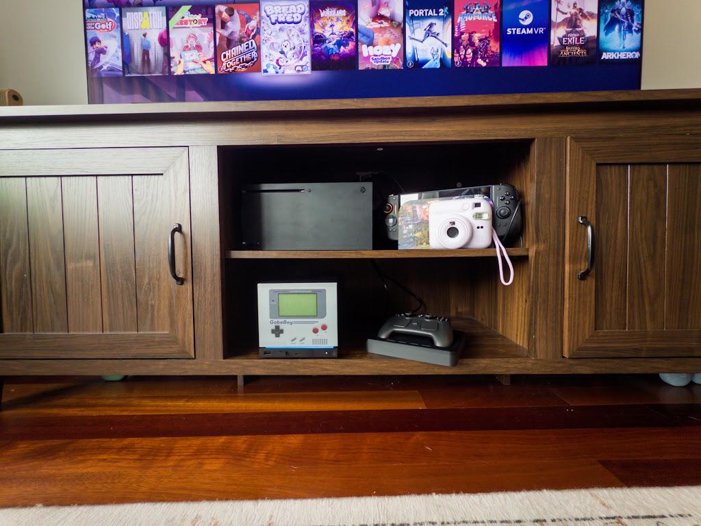 GabeBoy Retro Handheld Style Steam Machine Faceplate on the console, view of the entertainment console shelf with the console beside a game controller and a TV showing game covers above