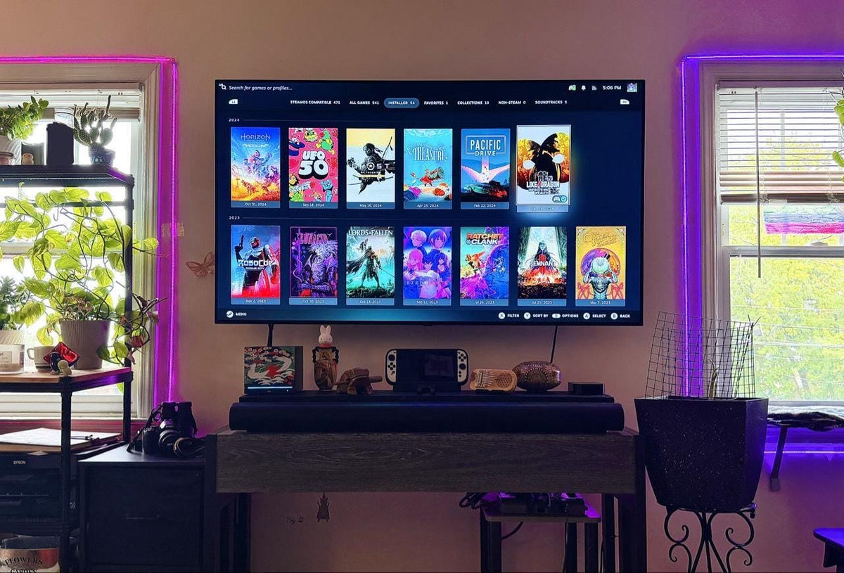 Japanese Crane Steam Machine Faceplate – UV Printed Front Panel | Red Sun & Cherry Blossom Art | Custom Mini PC Cover | Gamer Gift, wide room view of the crane faceplate installed on the console on a TV stand below a large wall-mounted TV showing a game library, with purple ambient lighting, windows, and plants nearby
