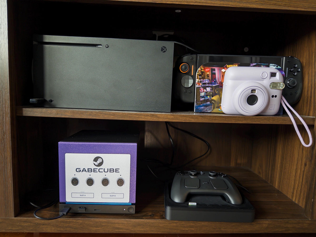 3D Printed GameCube Faceplate – Indigo Purple Steam Machine Controller Panel, GabeCube faceplate installed on the console on the lower shelf of a wooden media cabinet beside a Steam controller on its dock, with an Xbox, instant photo print and white instant camera on the shelf above