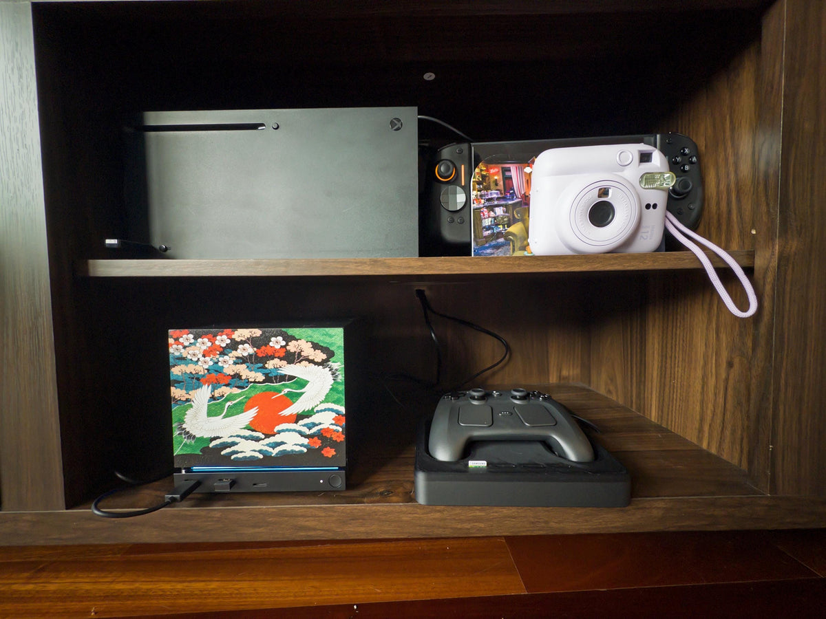 Japanese Crane Steam Machine Faceplate – UV Printed Front Panel | Red Sun & Cherry Blossom Art | Custom Mini PC Cover | Gamer Gift, crane faceplate installed on the lower shelf of a wooden media cabinet beside a Steam controller on its dock, with an Xbox, instant photo print, and white instant camera on the shelf above