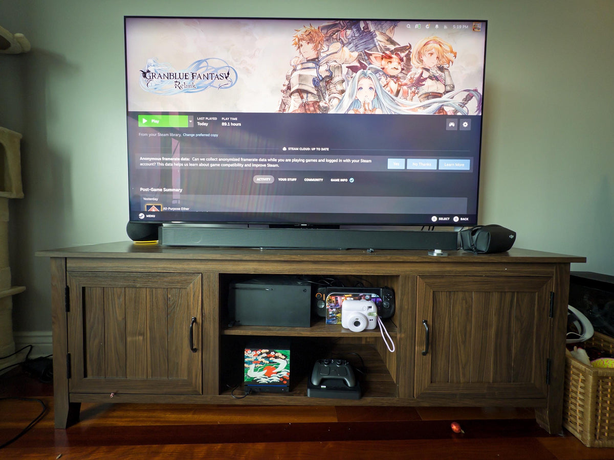 Japanese Crane Steam Machine Faceplate – UV Printed Front Panel | Red Sun & Cherry Blossom Art | Custom Mini PC Cover | Gamer Gift, wide view of a wooden media cabinet with a television showing a video game screen on top, the console with the crane faceplate installed on the lower open shelf next to a Steam controller on its dock