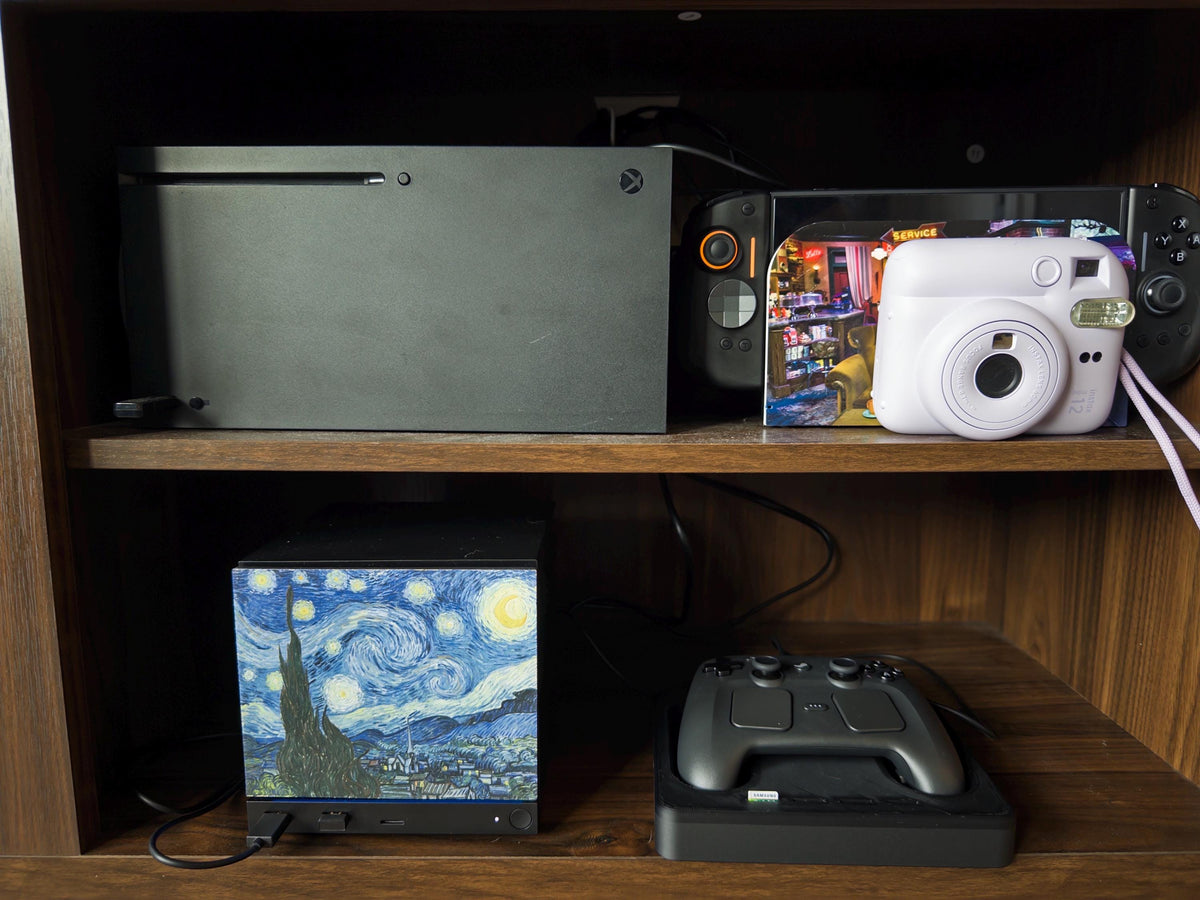 Starry Night Steam Machine Faceplate: Van Gogh Art UV Printed Magnetic Panel, Steam Machine with the Starry Night faceplate installed on the lower shelf of a wooden shelf, beside a black controller on its charging dock, with an Xbox, white instant camera and Steam Deck on the shelf above