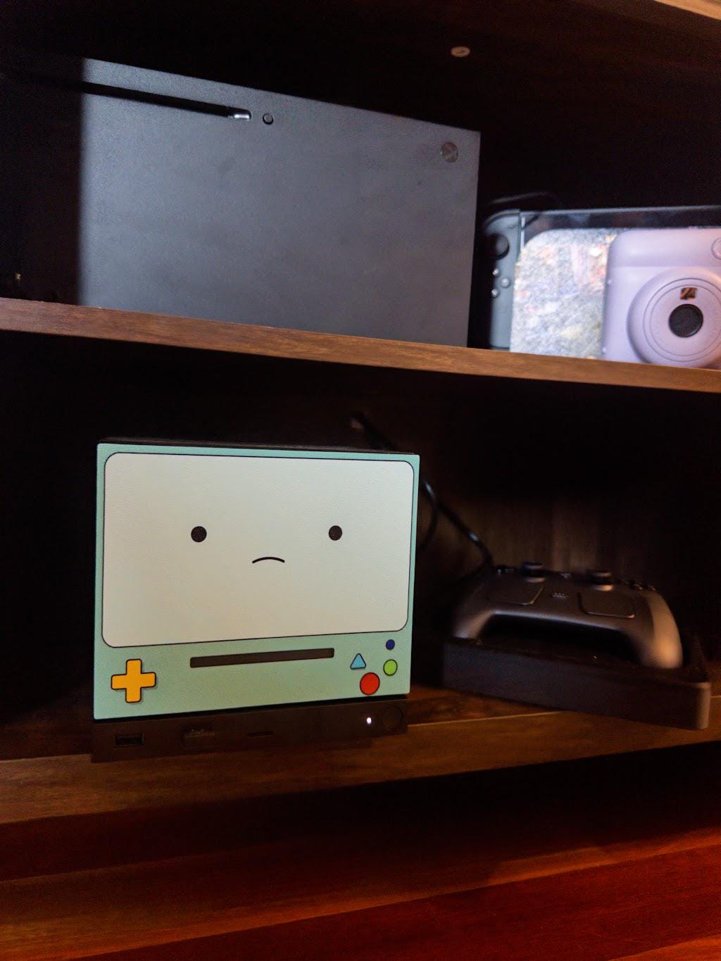 Robot Face Console Skin 2-Pack, Happy Sad Expression, Kawaii Gaming Setup Upgrade, sad expression skin installed on the console on a wooden shelf beside a game controller, with a white instant camera and other electronics on the shelf above