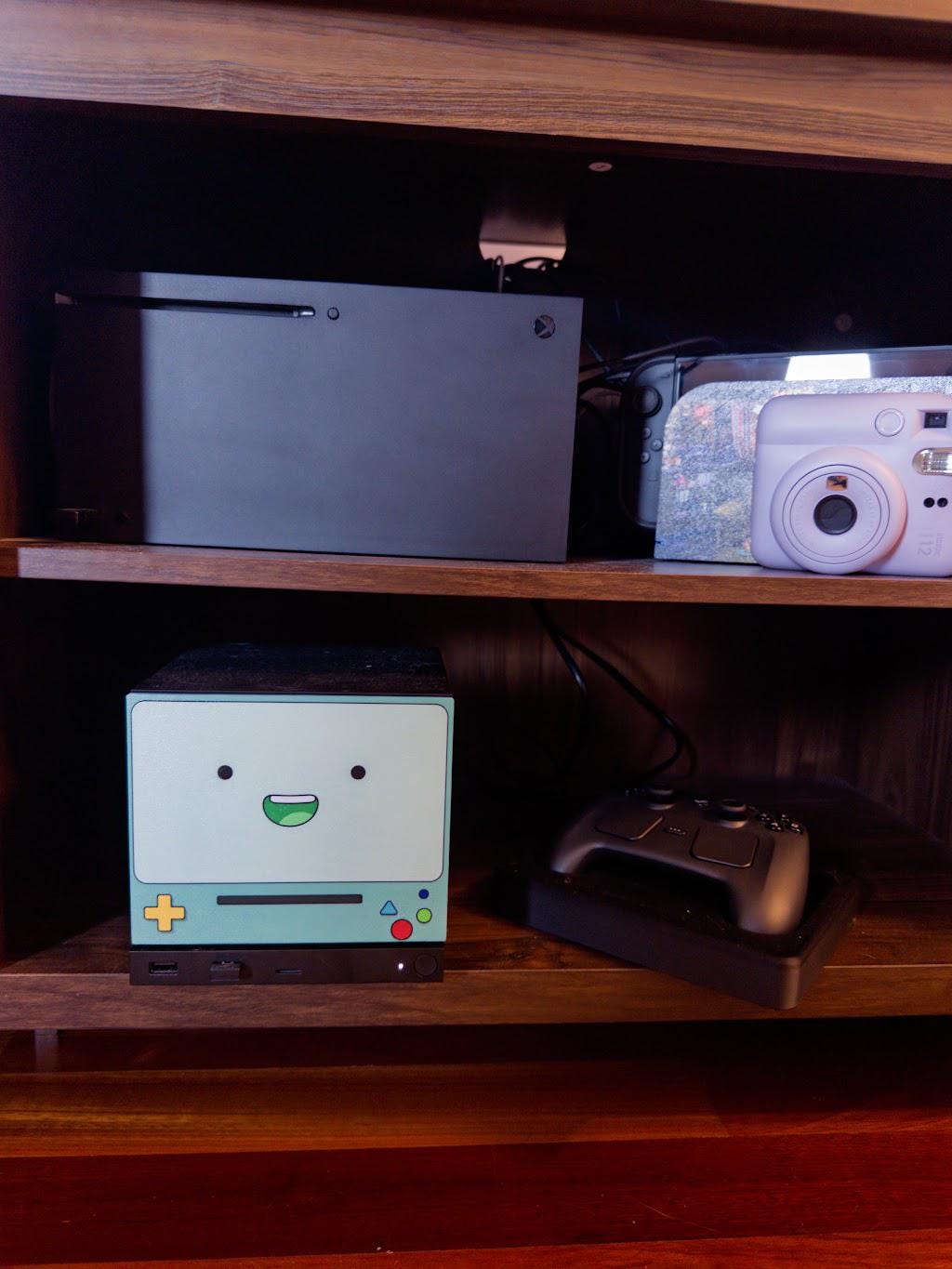 Robot Face Console Skin 2-Pack, Happy Sad Expression, Kawaii Gaming Setup Upgrade, happy expression skin shown installed on the console on a wooden shelf next to a game controller, with other consoles and a white instant camera on the shelf above