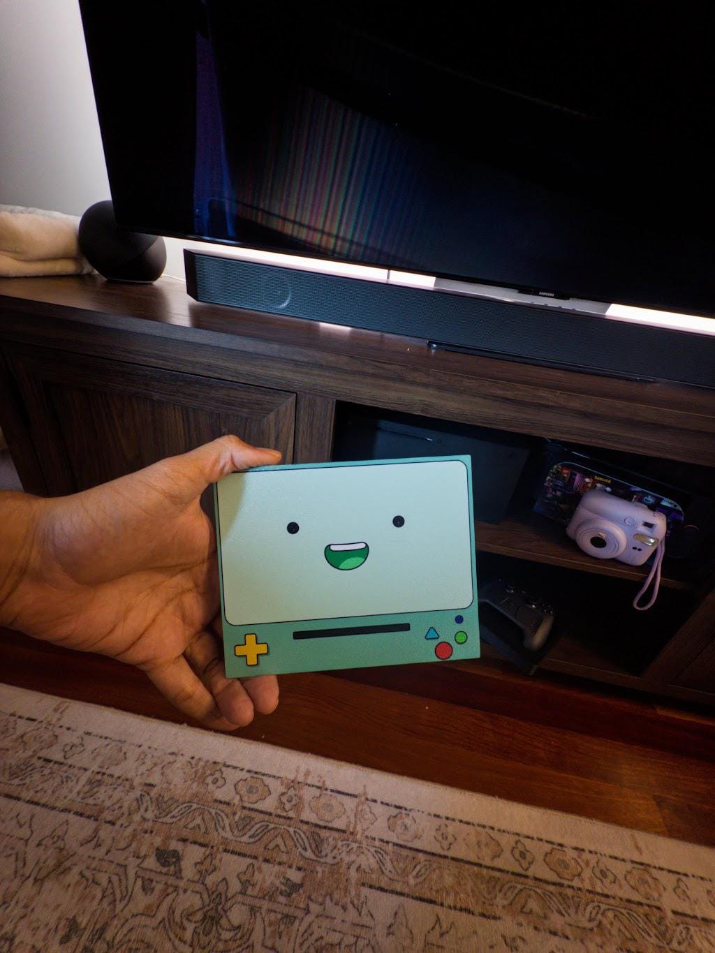 Robot Face Console Skin 2-Pack, Happy Sad Expression, Kawaii Gaming Setup Upgrade, happy grinning expression skin plate held in a hand showing the printed robot face front, in front of a TV stand with a television and soundbar