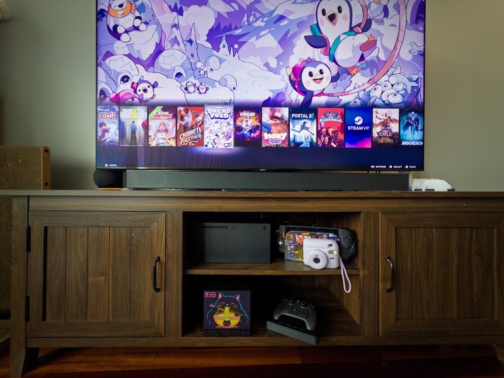 Steam Machine Faceplate Neon Ramen Cat Art | Magnetic UV Printed Panel | Kawaii Japanese Gamer Decor, wide shot of the Neon Ramen Cat faceplate installed on the console in a dark wood media console shelf below a large TV showing a game library, with a game controller and cameras nearby
