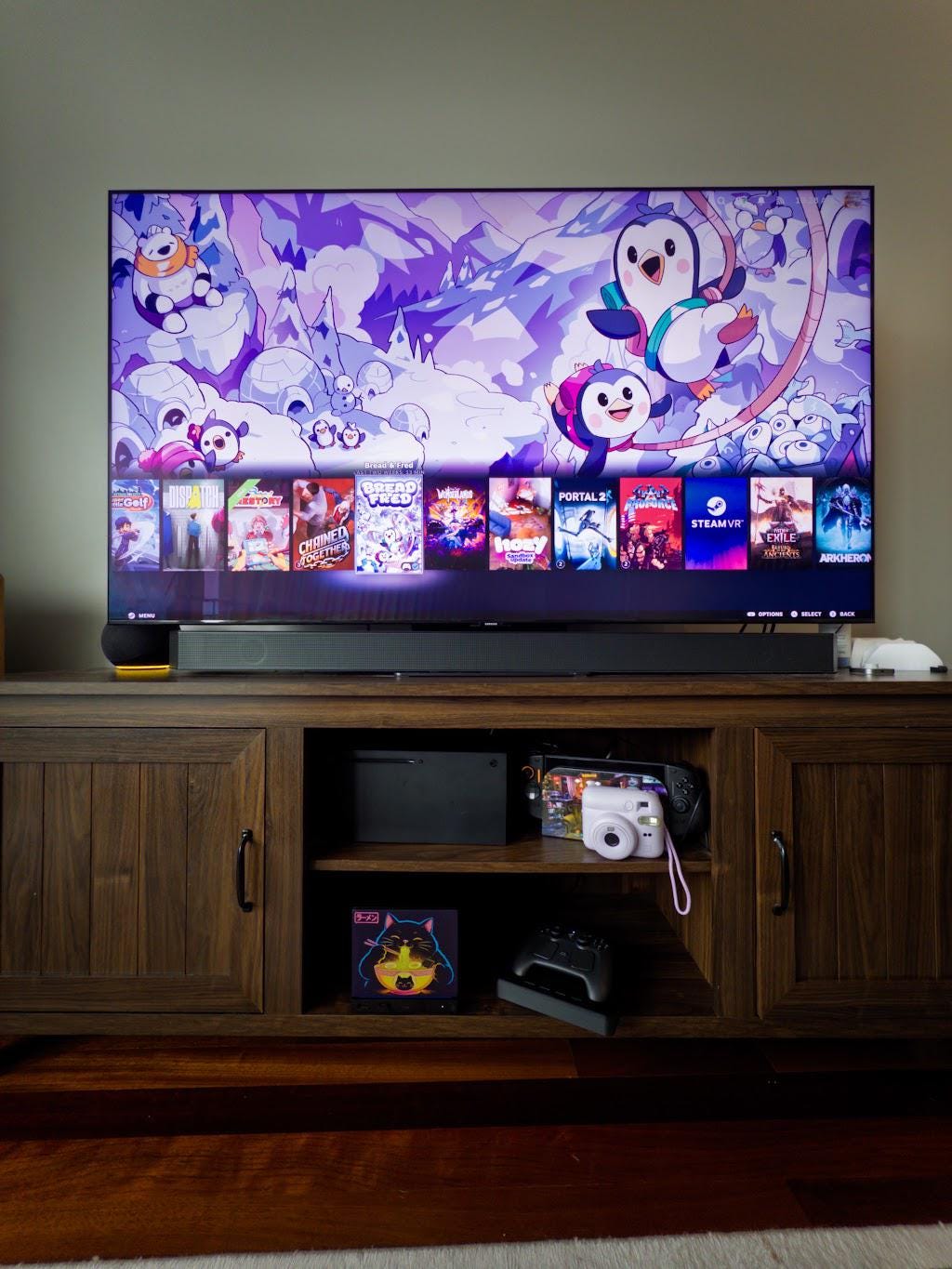 Steam Machine Faceplate Neon Ramen Cat Art | Magnetic UV Printed Panel | Kawaii Japanese Gamer Decor, wide view of the Neon Ramen Cat faceplate installed on the console inside a dark wood media console below a large TV, with a game controller and instant camera nearby