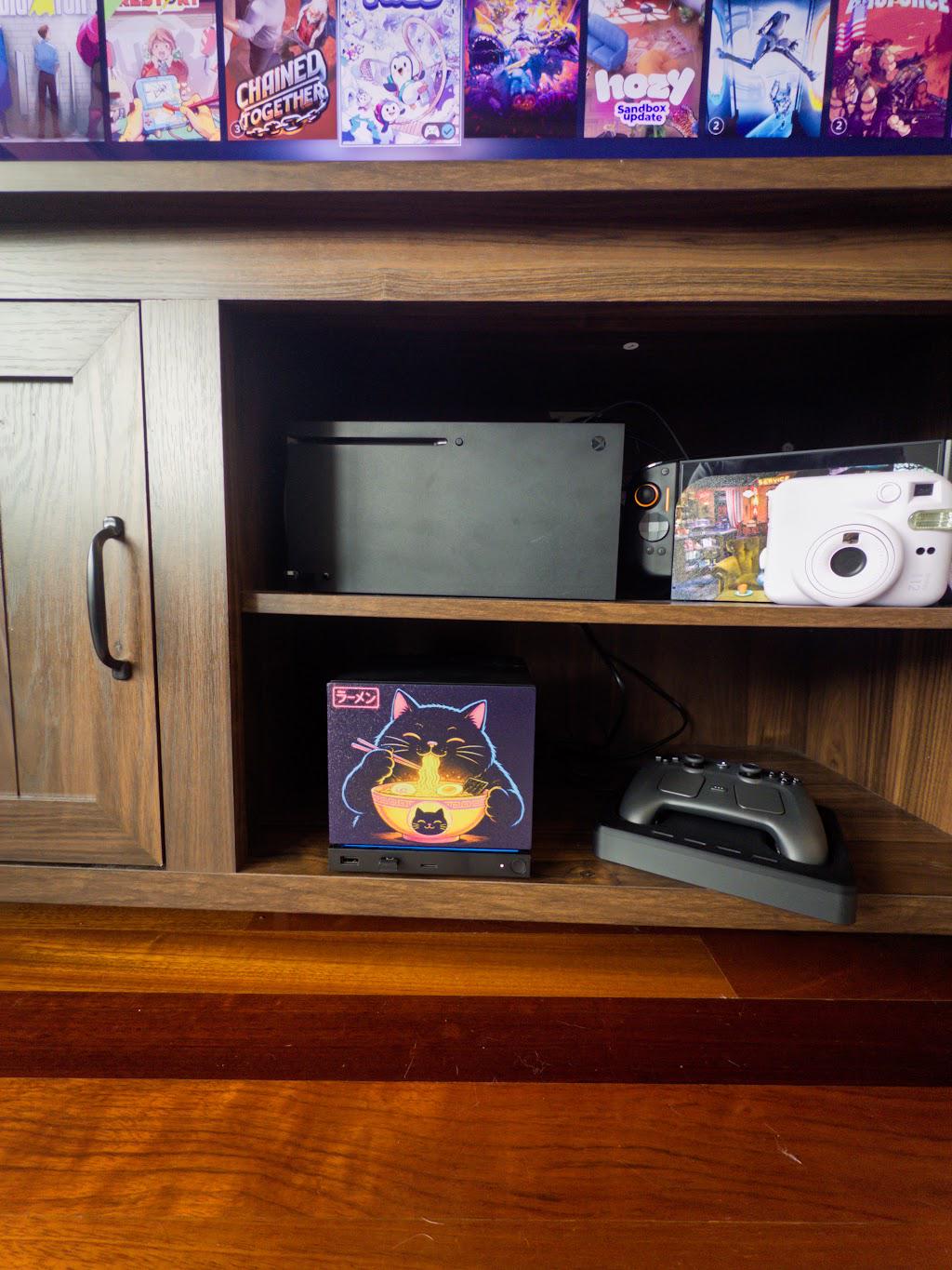 Steam Machine Faceplate Neon Ramen Cat Art | Magnetic UV Printed Panel | Kawaii Japanese Gamer Decor, Neon Ramen Cat faceplate installed on the console in a wooden media console shelf beside a Steam controller on its dock, with a game console and instant camera on the shelf above