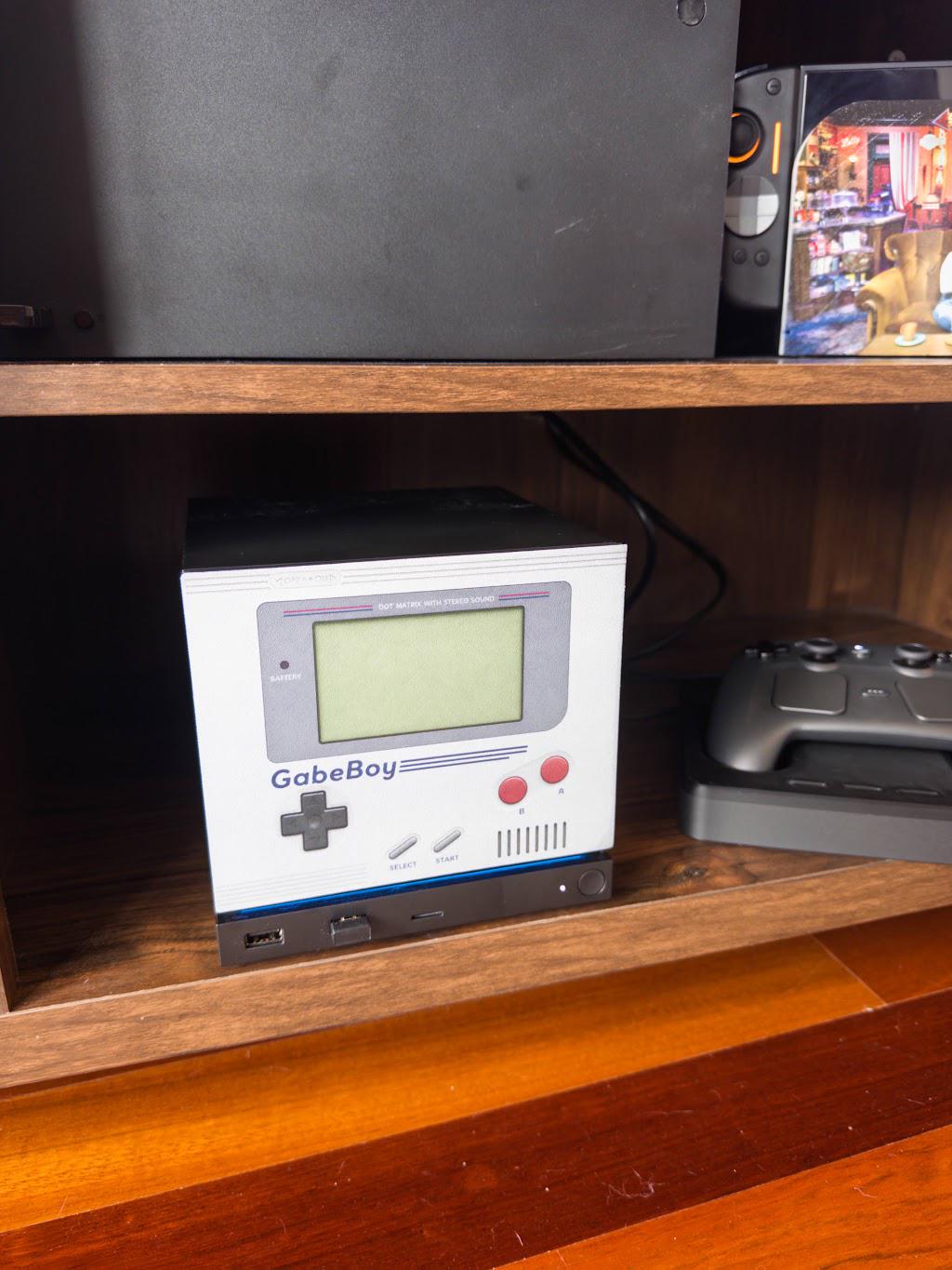 GabeBoy Retro Handheld Style Steam Machine Faceplate on the console, styled on a wooden entertainment shelf beside a game controller