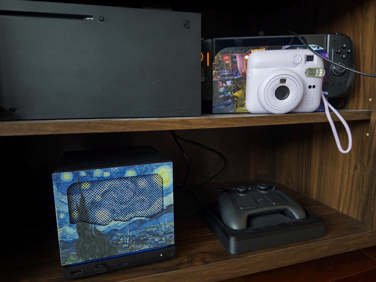 New Mesh!! Starry Night Steam Machine Faceplate Van Gogh Front Panel Fine Art UV Printed Magnetic Classic Painting Gamer Gift on the console on the lower wooden shelf next to a game controller, with a white instant camera and other consoles on the upper shelf