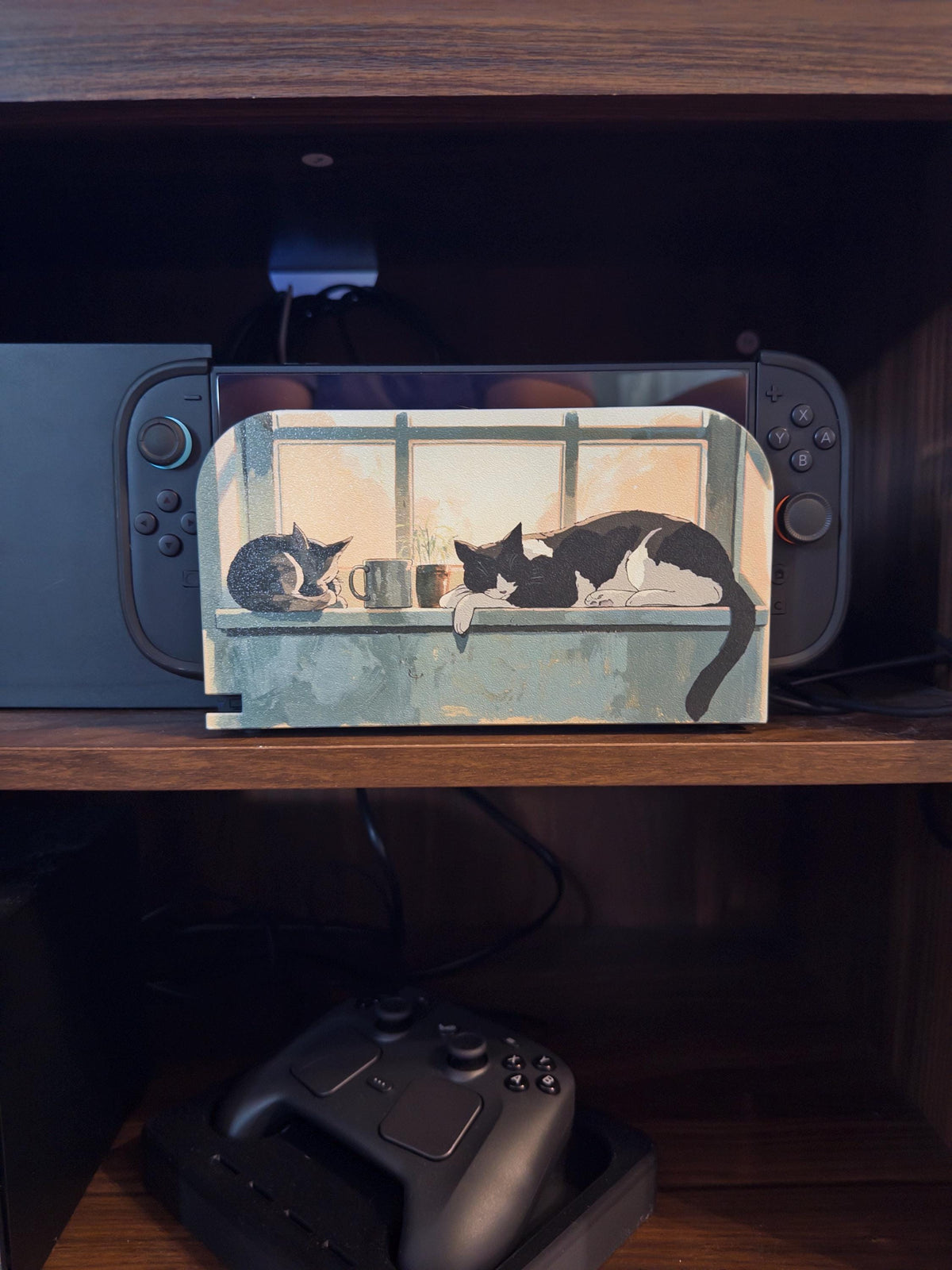 Custom 3D Printed Switch 2 Dock Faceplate, Magnetic Snap-On Cover (Personalized Photo), close-up of the dock with the two-cats-by-a-window faceplate installed on a wooden shelf, with the console behind it and a game controller on a charging dock below