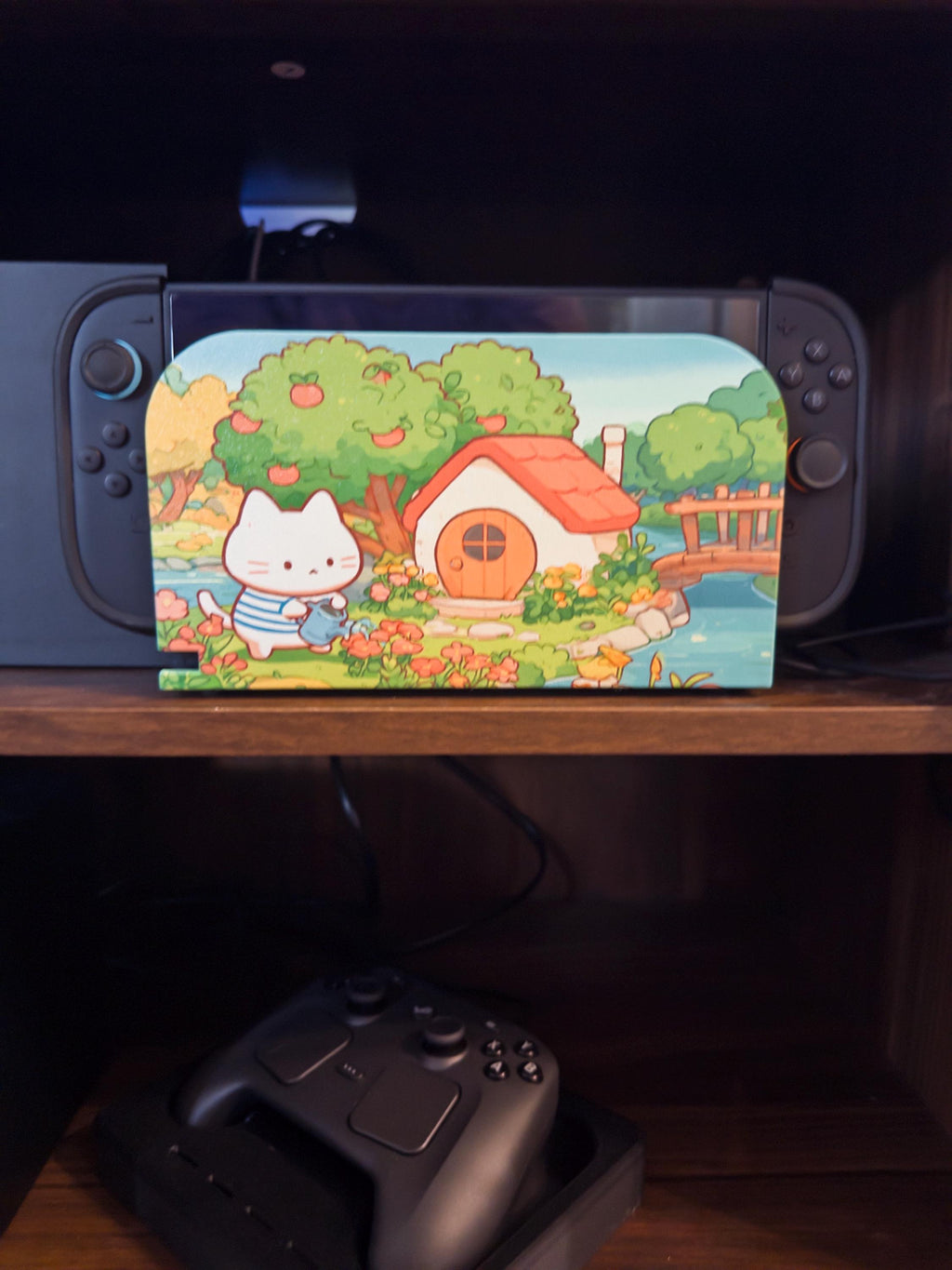 Custom 3D Printed Switch 2 Dock Faceplate, Magnetic Snap-On Cover (Personalized Photo), close-up of the dock with the white cat watering flowers cottage faceplate installed on a wooden shelf, with a game controller on the shelf below