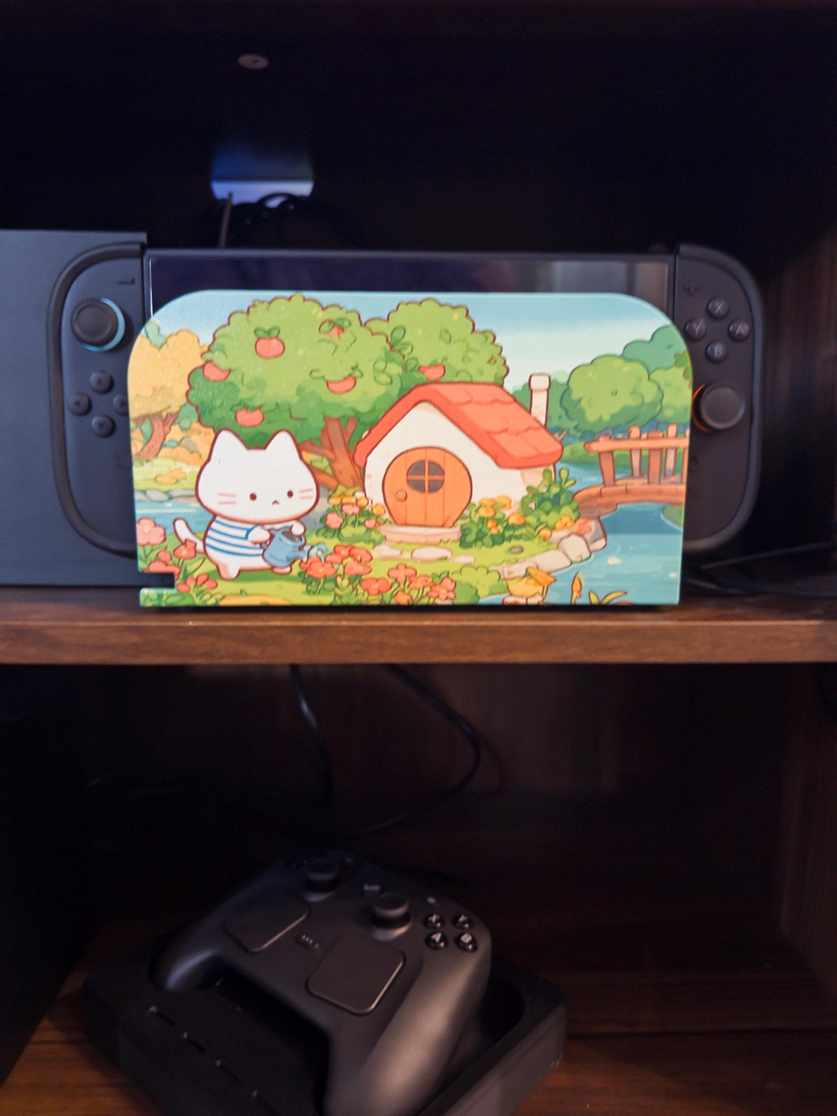 Custom 3D Printed Switch 2 Dock Faceplate, Magnetic Snap-On Cover (Personalized Photo), close-up of the dock with the white cat watering flowers cottage faceplate installed on a wooden shelf, with a game controller on the shelf below