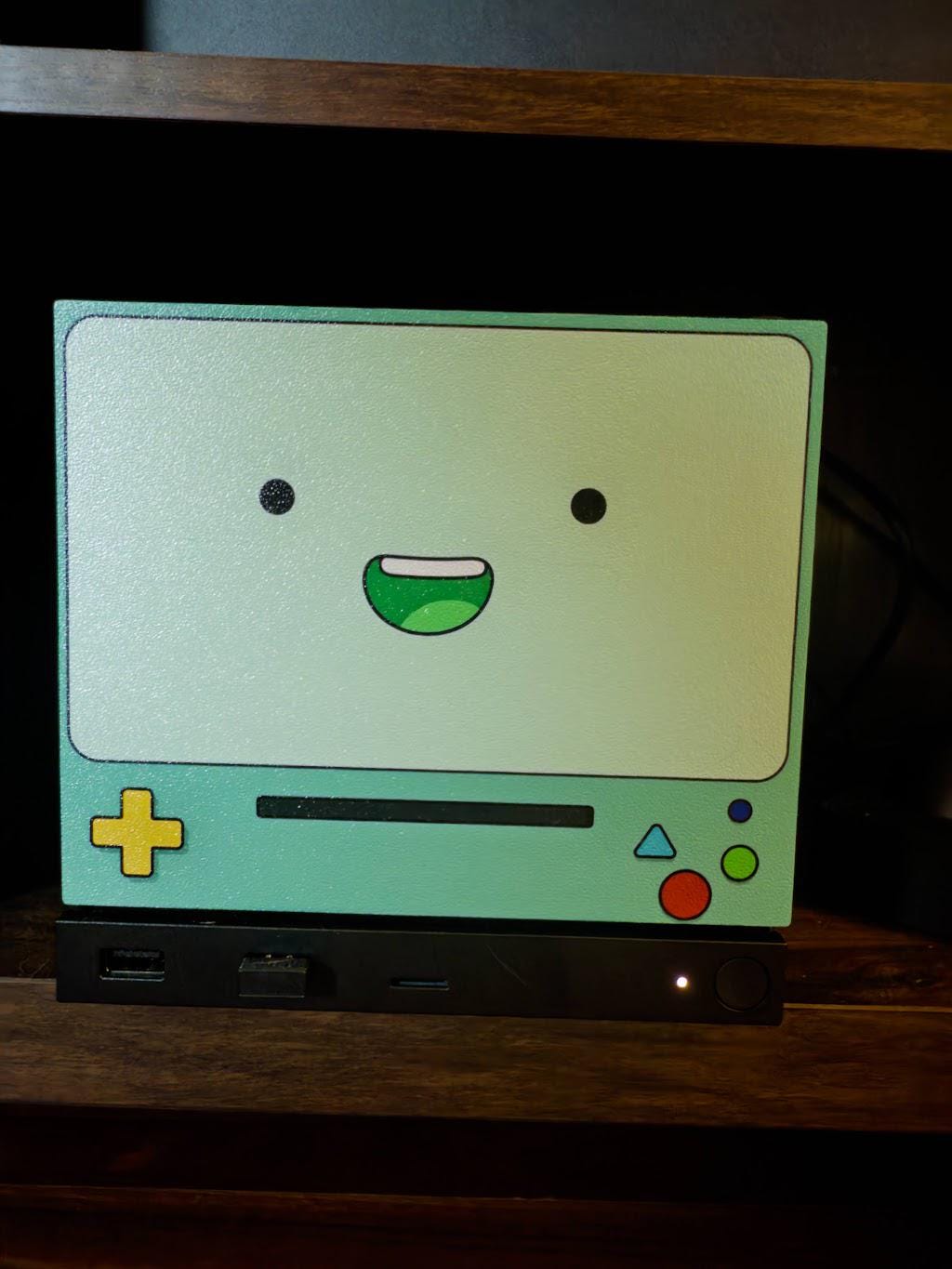 Robot Face Console Skin 2-Pack, Happy Sad Expression, Kawaii Gaming Setup Upgrade, happy grinning expression skin shown installed on the console in a close-up front view on a dark wooden shelf