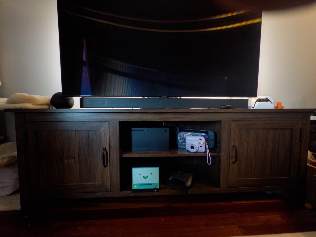 Robot Face Console Skin 2-Pack, Happy Sad Expression, Kawaii Gaming Setup Upgrade, happy expression skin installed on the console visible on the lower shelf of a dark wood media console, wide living room shot with a TV, soundbar and other electronics