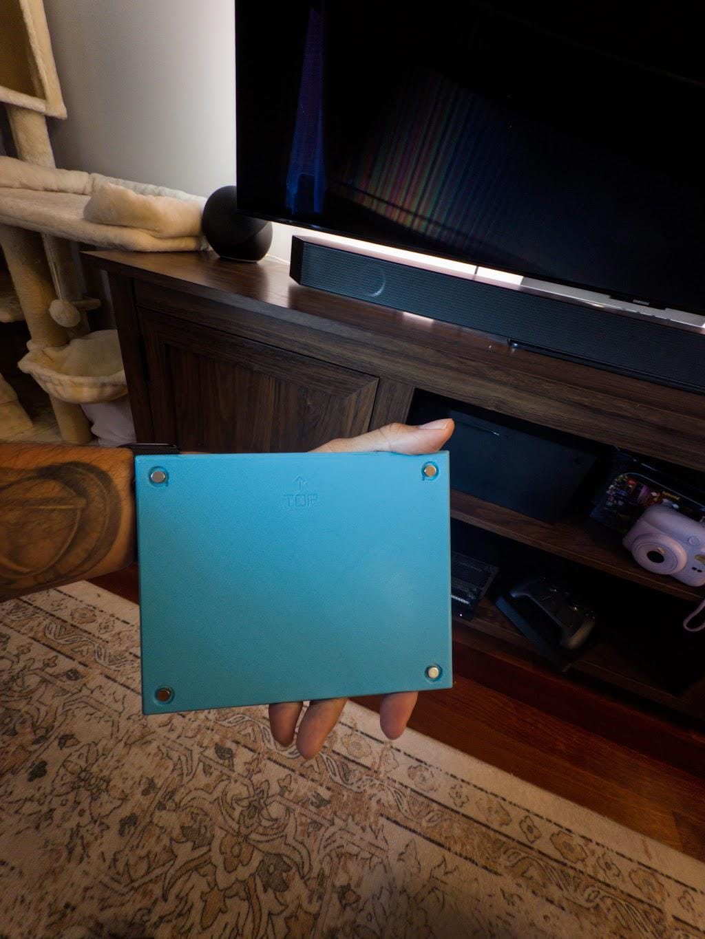Robot Face Console Skin 2-Pack, Happy Sad Expression, Kawaii Gaming Setup Upgrade, back of a light blue skin plate held in a hand showing the corner mounting screws and TOP marking, with a dark wood TV stand and television in the background
