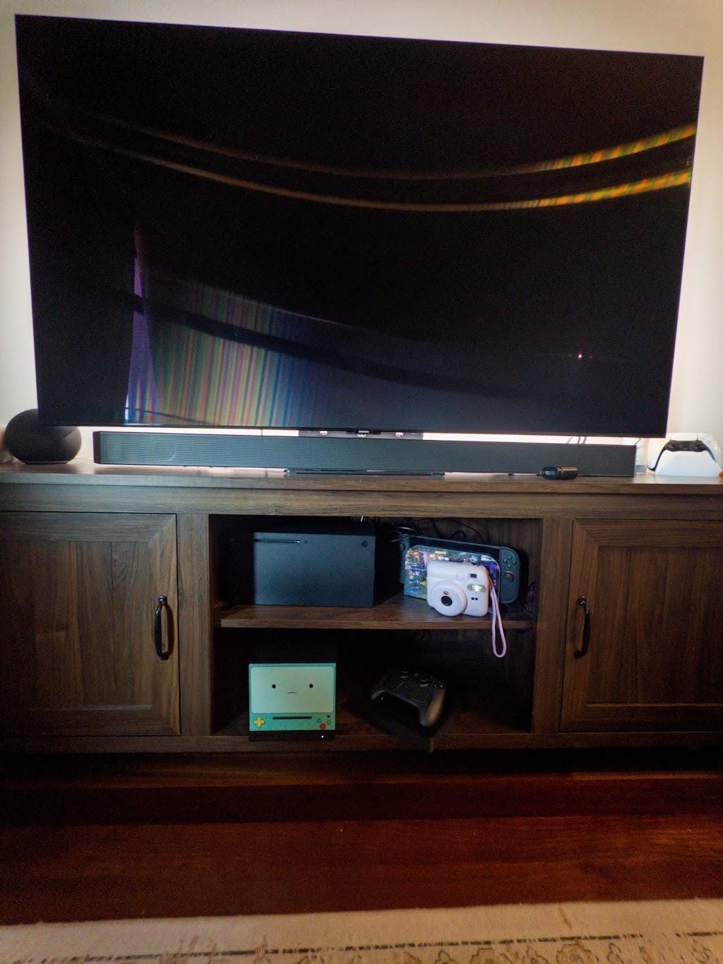 Robot Face Console Skin 2-Pack, Happy Sad Expression, Kawaii Gaming Setup Upgrade, happy expression skin visible on the console on the middle shelf of a wooden TV stand, wide living room view with a large television and soundbar above