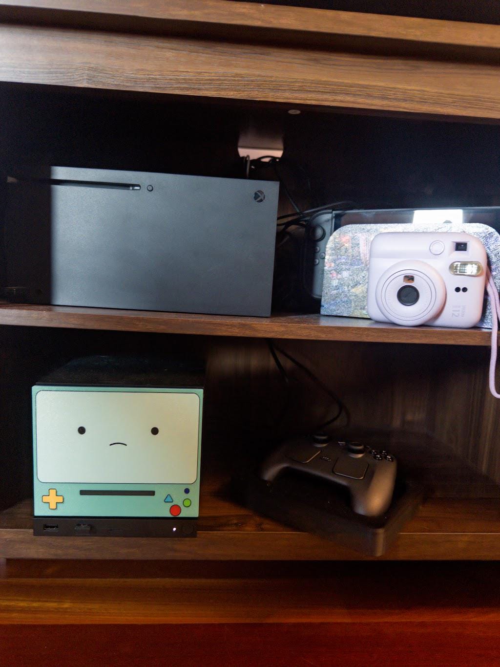 Robot Face Console Skin 2-Pack, Happy Sad Expression, Kawaii Gaming Setup Upgrade, sad expression skin installed on the console on a wooden shelf beside a game controller, with a black game console, white instant camera and other electronics on the shelf above