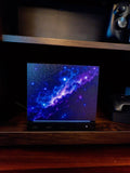 Galaxy Steam Machine Faceplate, UV Printed Nebula Cover, Custom Valve Console Accessory with the purple and blue galaxy nebula artwork, shown installed on the console on a dark wooden shelf with a game controller above