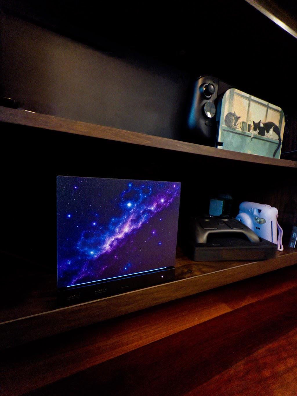Galaxy Steam Machine Faceplate, UV Printed Nebula Cover, Custom Valve Console Accessory with the purple and blue galaxy nebula artwork, angled view installed on the console on a dark wooden shelf with a game controller and an instant camera nearby