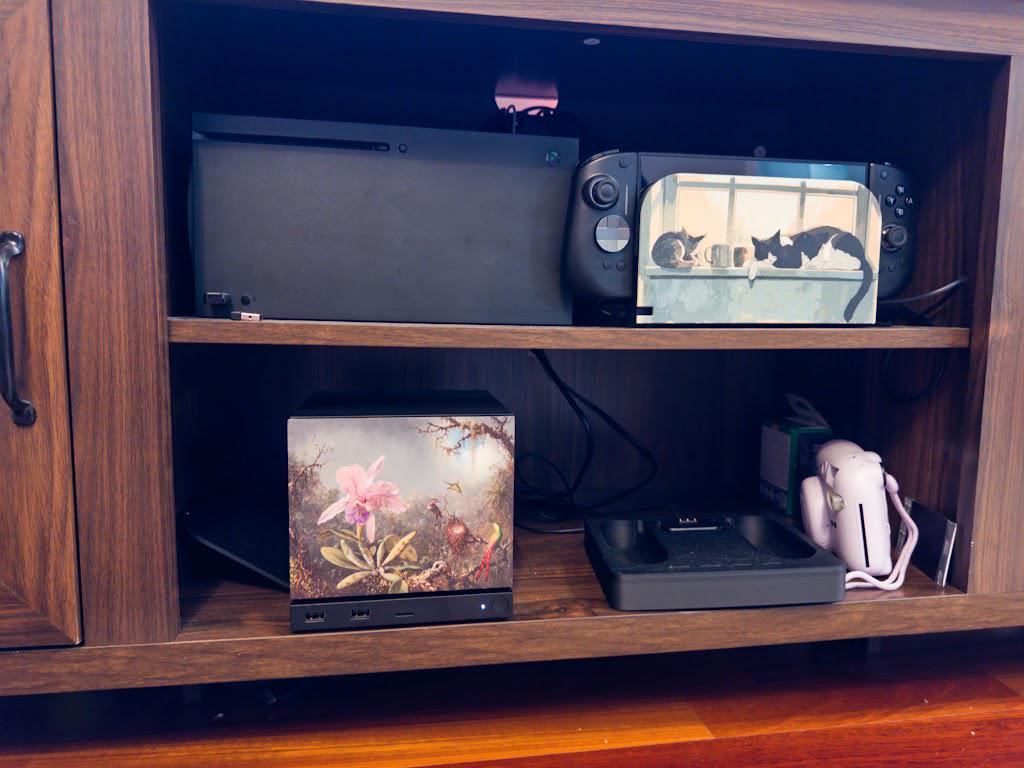 Steam Machine Faceplate, Hummingbird Orchid Botanical Art Decor, Vintage Gamer Accessory on the console, wide shot on the lower shelf of a wooden entertainment cabinet beside a white instant camera and a charging dock