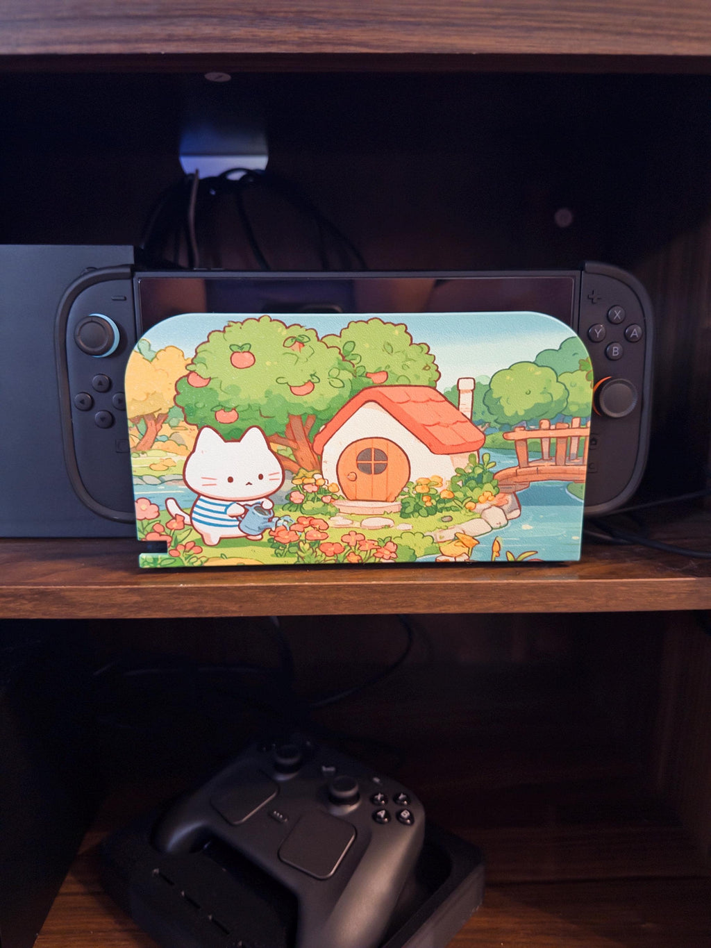 Custom 3D Printed Switch 2 Dock Faceplate, Magnetic Snap-On Cover (Personalized Photo), faceplate with a cartoon illustration of a white cat watering flowers beside a cottage with apple trees and a pond, shown installed on the Switch 2 dock on a wooden shelf
