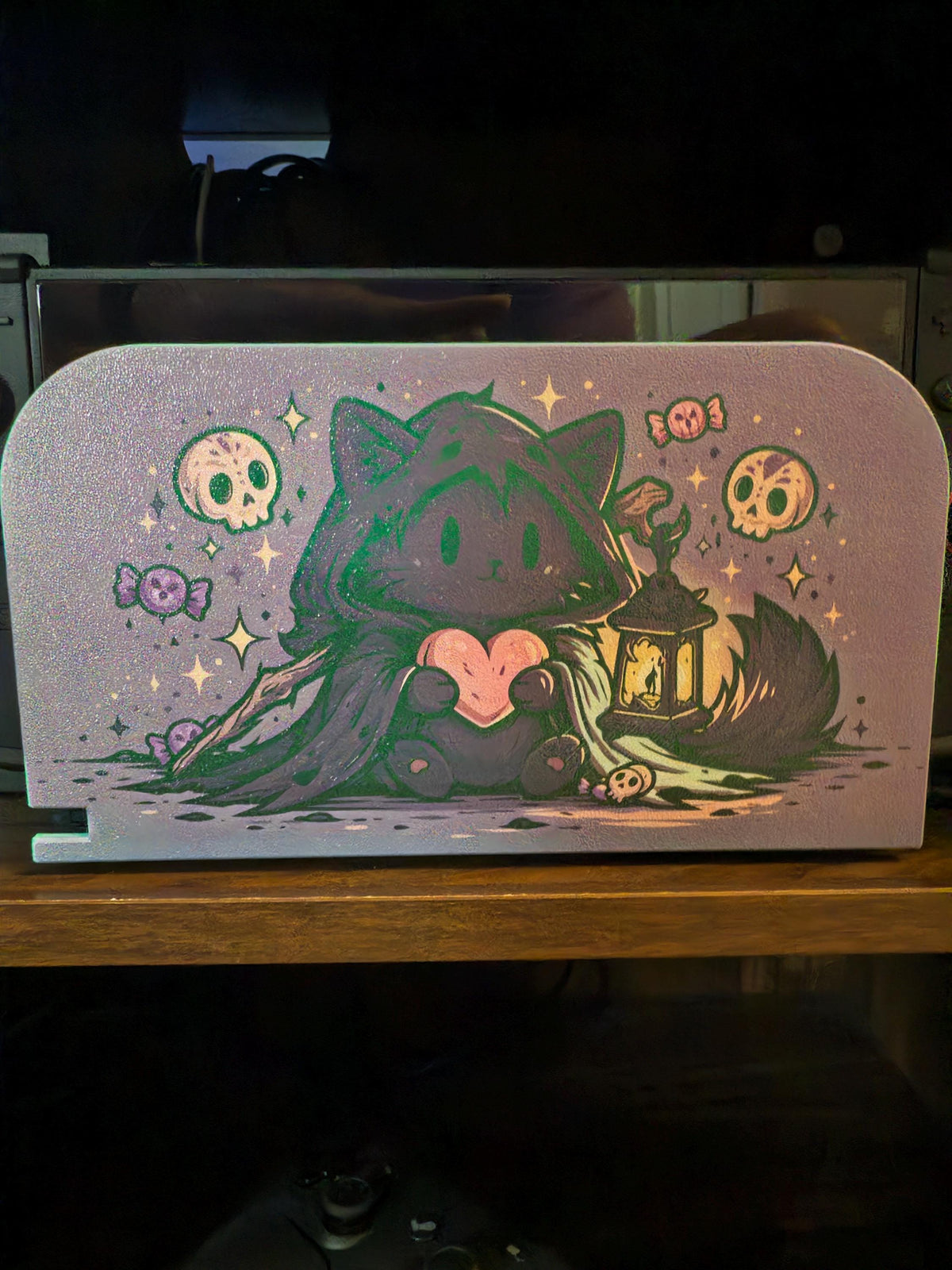 Custom 3D Printed Switch 2 Dock Faceplate, Magnetic Snap-On Cover (Personalized Photo), faceplate with an illustration of a hooded black cat holding a pink heart surrounded by skulls, stars, candies and a lantern, shown installed on the Switch 2 dock on a wooden shelf