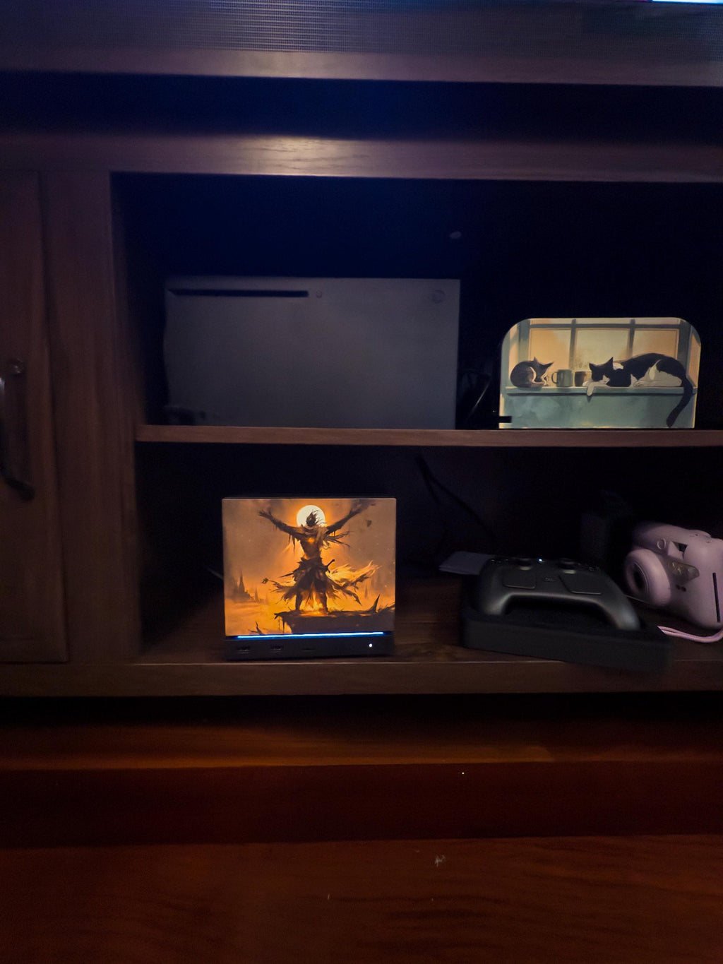 Praise the Sun Steam Machine Faceplate – Dark Fantasy Silhouette (Magnetic UV Print) with the fiery warrior artwork, shown installed on the console on a wooden entertainment shelf beside a game controller, with a PlayStation 5 and other electronics on the shelf above