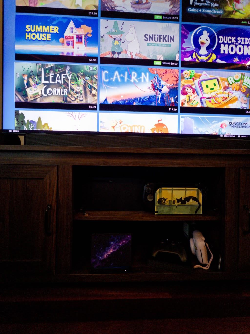 Galaxy Steam Machine Faceplate, UV Printed Nebula Cover, Custom Valve Console Accessory with the galaxy nebula artwork, seen on the lower shelf of a wooden media console beneath a TV showing a game library, with a game controller beside it