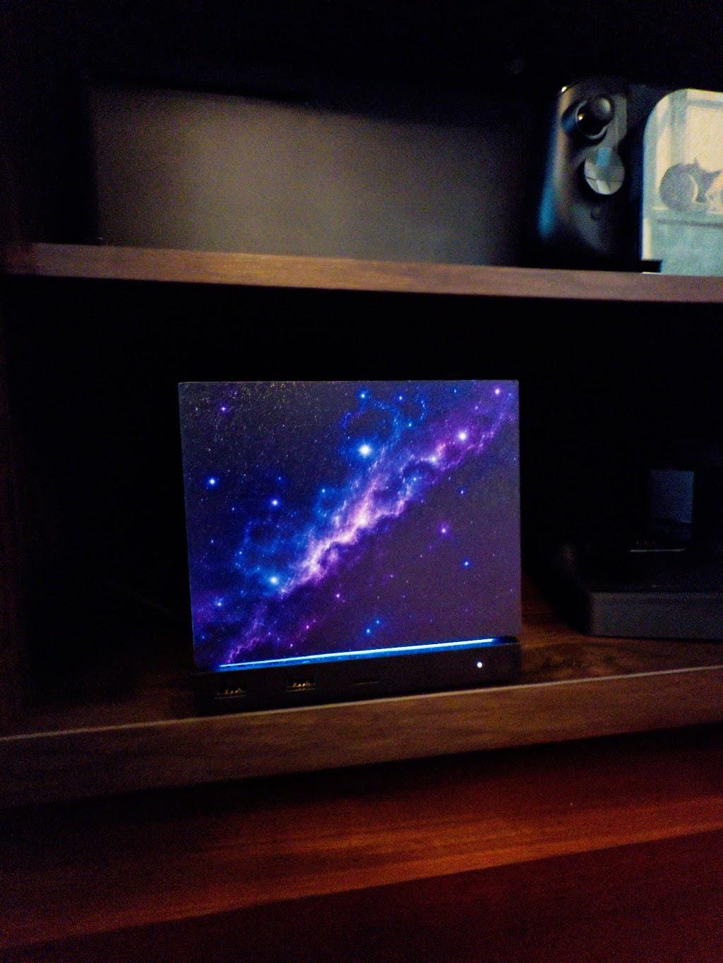 Galaxy Steam Machine Faceplate, UV Printed Nebula Cover, Custom Valve Console Accessory with the purple and blue galaxy nebula artwork, close-up straight-on view installed on the console on a dark wooden shelf with a game controller on the shelf above