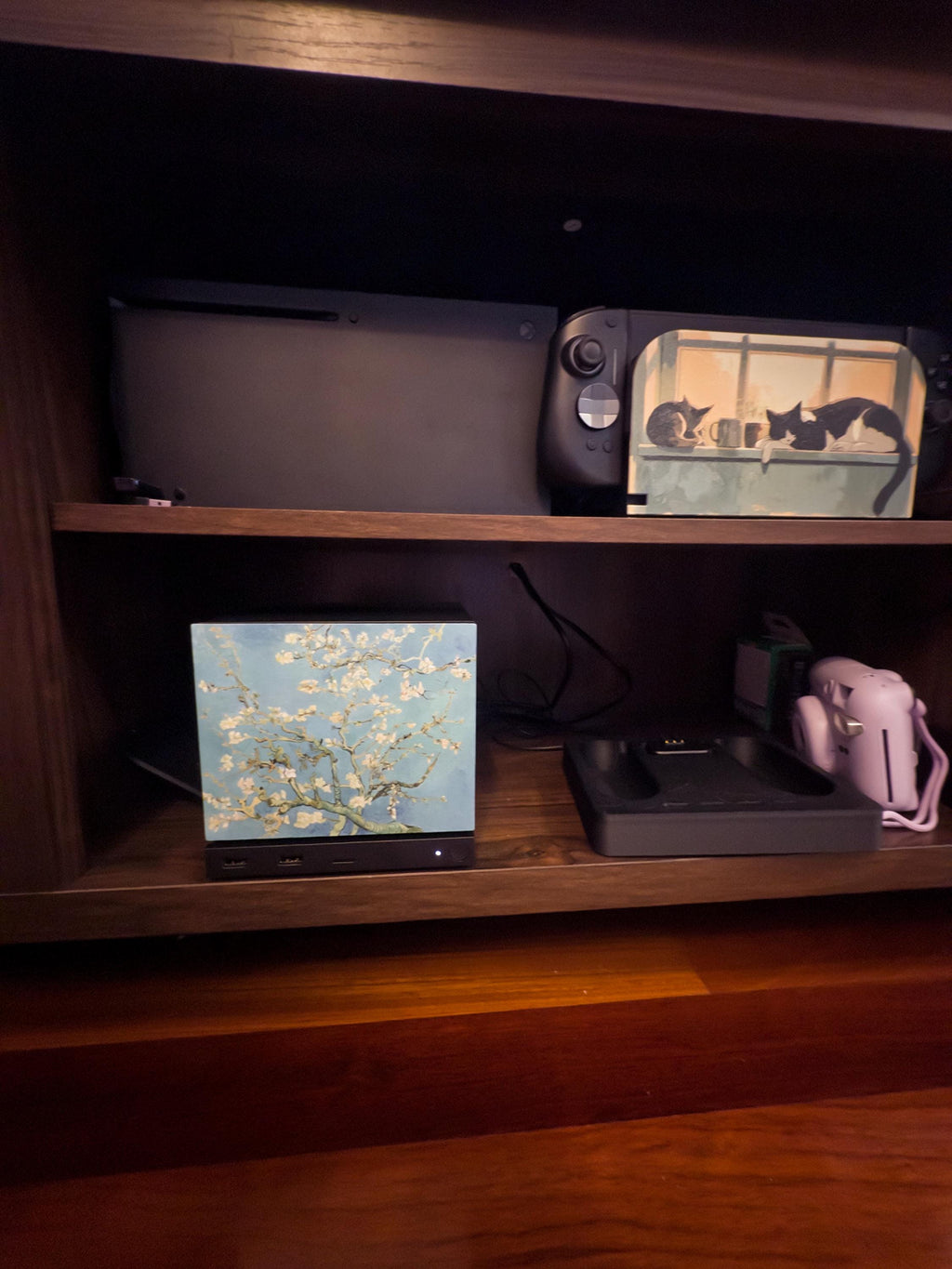 Van Gogh Almond Blossom Steam Machine Faceplate, Magnetic Art Cover Gaming Decor, wider shelf view of the Steam Machine with the Almond Blossom faceplate installed on the lower shelf of a dark wooden unit, with an Xbox and Steam Deck on the shelf above and an instant camera beside it