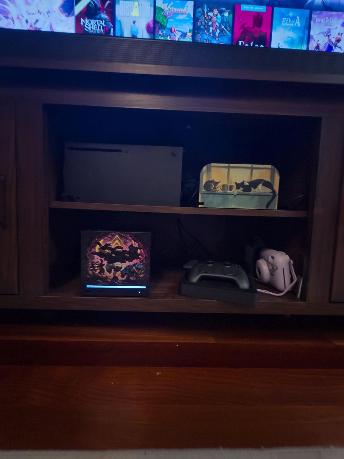 Dungeon Crawler Carl Steam Machine Faceplate | Licensed Landfin Studio Art (UV Printed) on the console, wide shot of a wooden media cabinet with the faceplate glowing beneath a large TV showing game covers, beside a game controller and a white instant camera