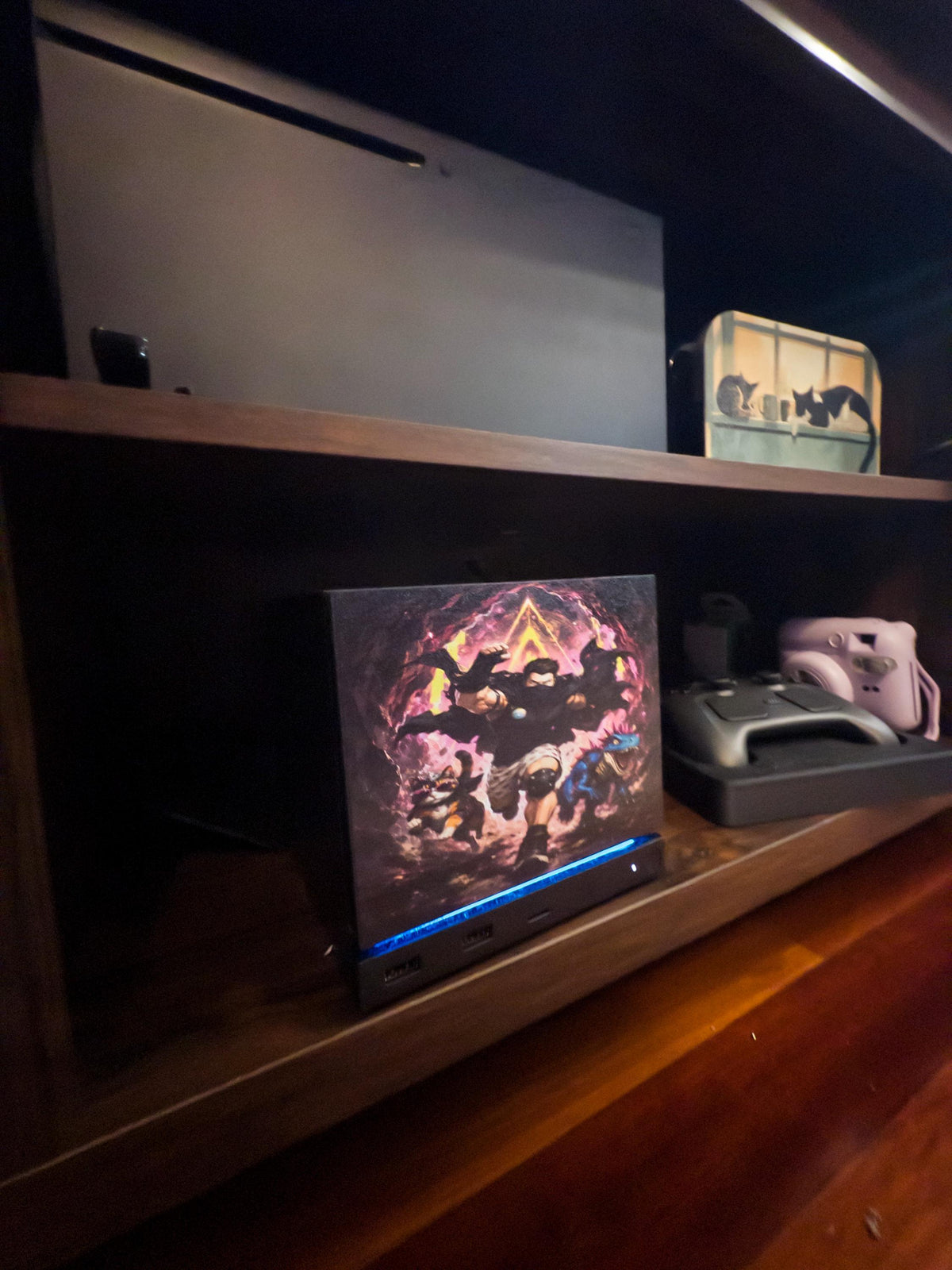 Dungeon Crawler Carl Steam Machine Faceplate | Licensed Landfin Studio Art (UV Printed) with Carl leaping through the glowing dungeon portal, angled view installed on the console on a wooden shelf beside a game controller and a white instant camera, with a larger console on the shelf above