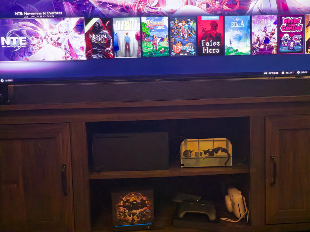 Praise the Sun Steam Machine Faceplate – Dark Fantasy Silhouette (Magnetic UV Print) with the fiery warrior artwork, visible on the console on the lower shelf of a wooden media cabinet beneath a large TV showing a game library interface