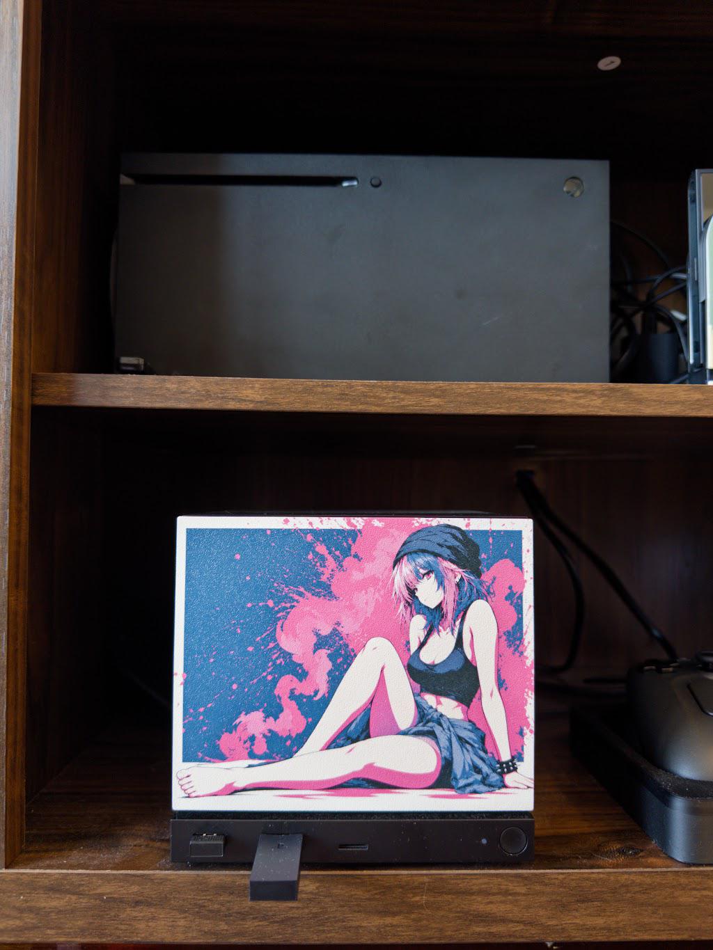 Anime Pinup Art Steam Machine Faceplate | Pink Blue Splatter Print | Magnetic UV Printed Panel | Stylish Gamer Gift with the pink and blue splatter anime girl art, frontal view of the faceplate installed on the console centered in a wooden shelf cubby, with a larger black game console on the shelf directly above