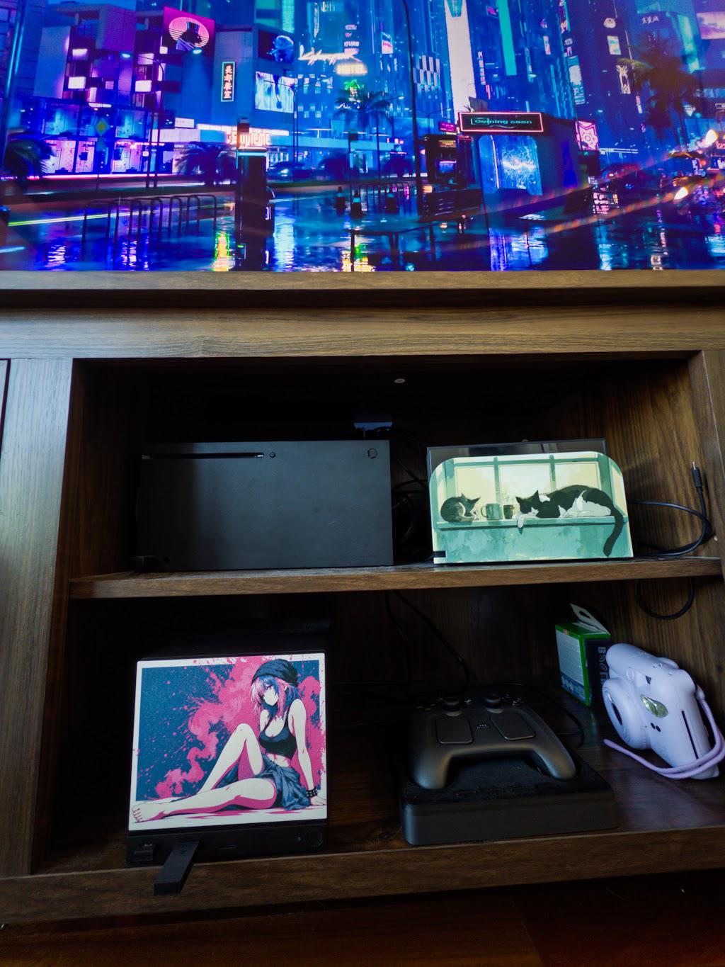 Anime Pinup Art Steam Machine Faceplate | Pink Blue Splatter Print | Magnetic UV Printed Panel | Stylish Gamer Gift with the pink and blue splatter anime girl art, shown installed on the console on the lower shelf of a wooden media cabinet beneath a TV displaying a neon city street scene, with a game controller and a white instant camera beside it and a small fish tank on the upper shelf