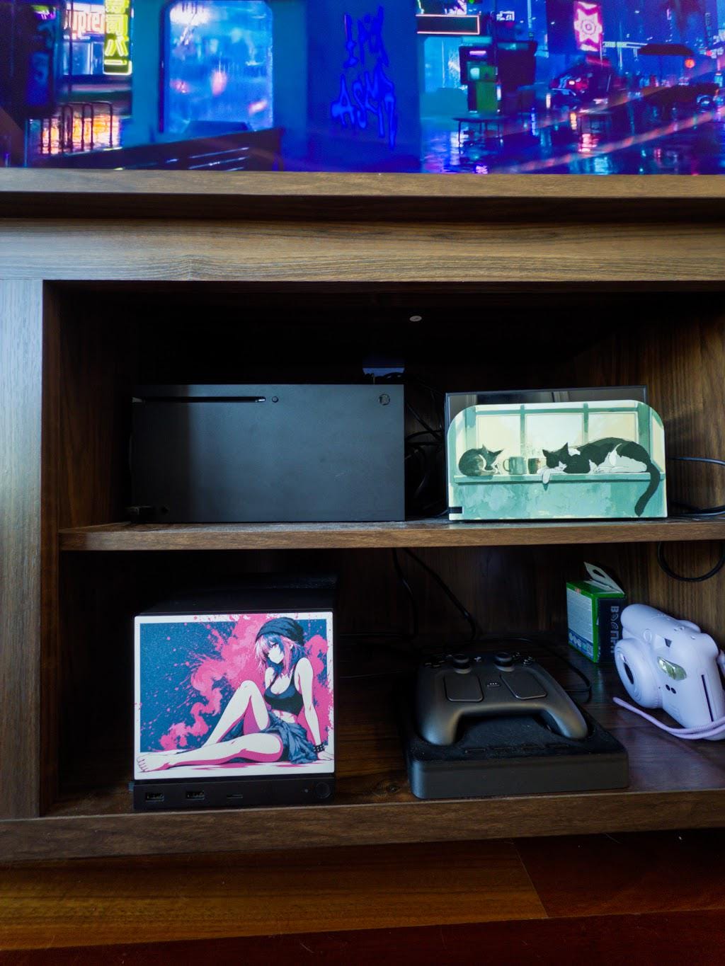 Anime Pinup Art Steam Machine Faceplate | Pink Blue Splatter Print | Magnetic UV Printed Panel | Stylish Gamer Gift with the pink and blue splatter anime girl art, visible on the console on the lower shelf of a wooden media cabinet beneath a TV showing a neon city game screen, beside a game controller and a white instant camera