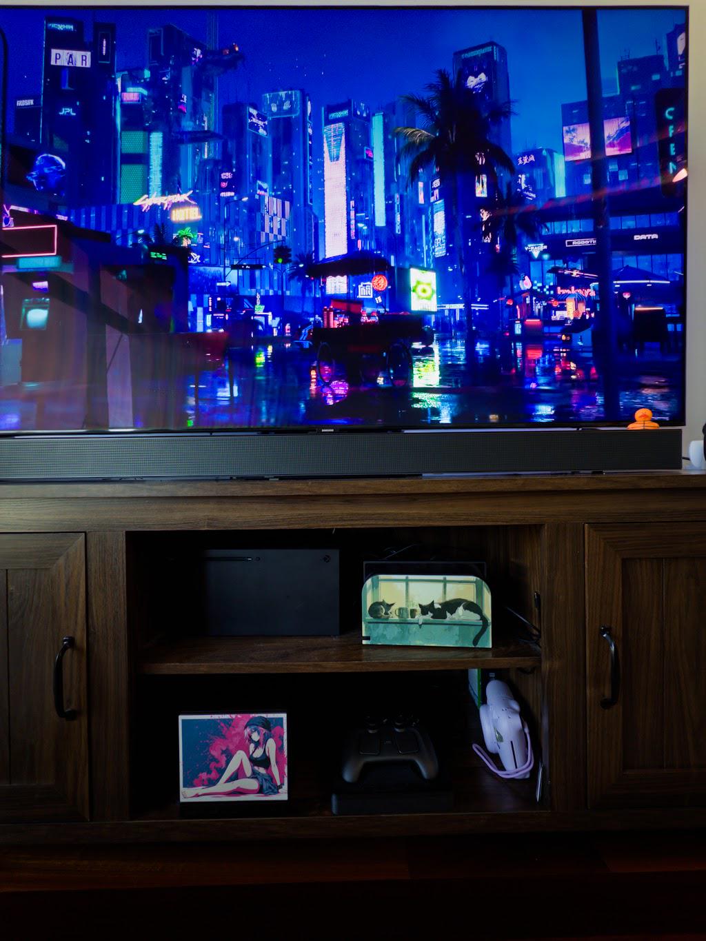 Anime Pinup Art Steam Machine Faceplate | Pink Blue Splatter Print | Magnetic UV Printed Panel | Stylish Gamer Gift with the pink and blue splatter anime girl art, visible on the console on the lower shelf of a wooden media cabinet beneath a large TV showing a neon cityscape, beside a game controller