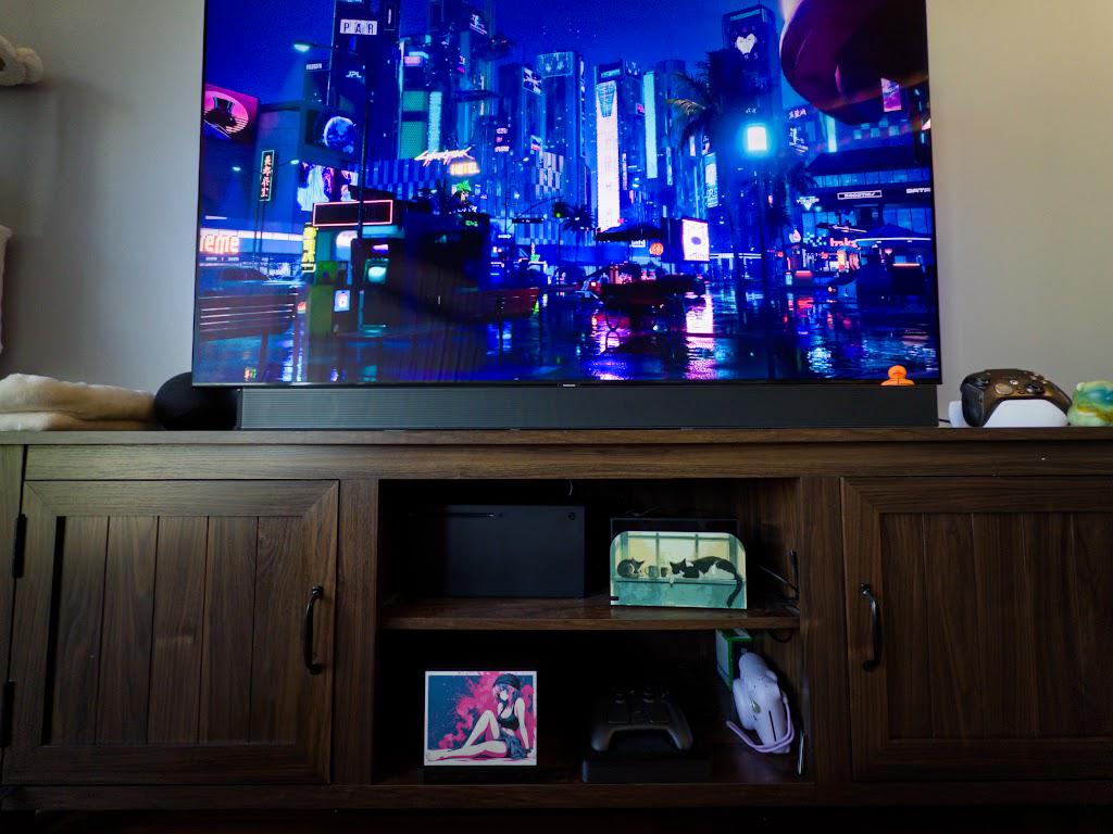 Anime Pinup Art Steam Machine Faceplate | Pink Blue Splatter Print | Magnetic UV Printed Panel | Stylish Gamer Gift with the pink and blue splatter anime girl art, visible on the console on the lower shelf of a wooden media console beneath a large TV showing a neon city scene, in a wide landscape shot beside a game controller