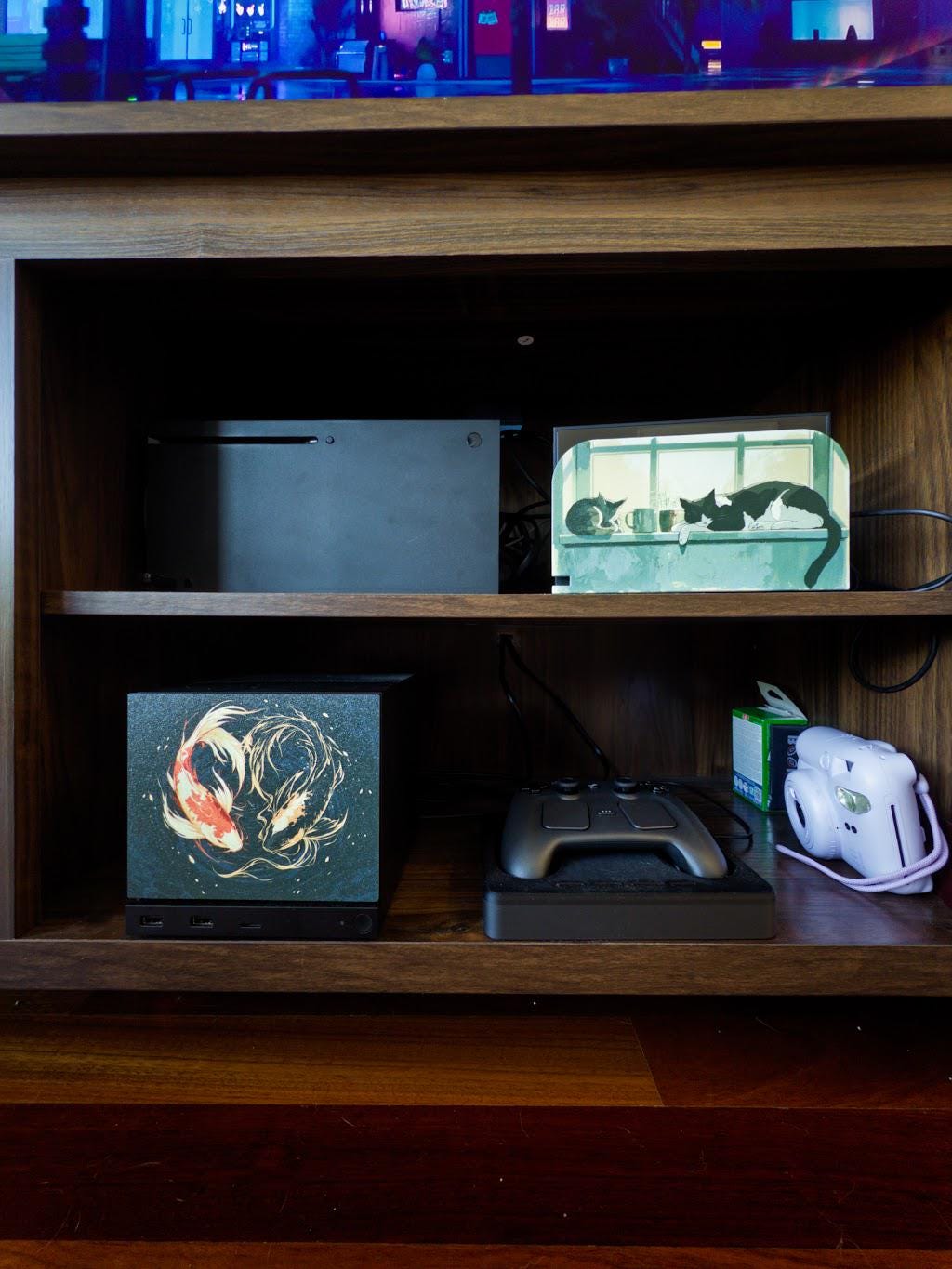 Koi Art Steam Machine Faceplate | Dark Teal Japanese Fish Panel | Magnetic UV Printed Cover | Zen Gamer Gift with two koi circling in dark teal water artwork, shown installed on the console on the lower shelf of a wooden media cabinet beside a game controller and a white instant camera