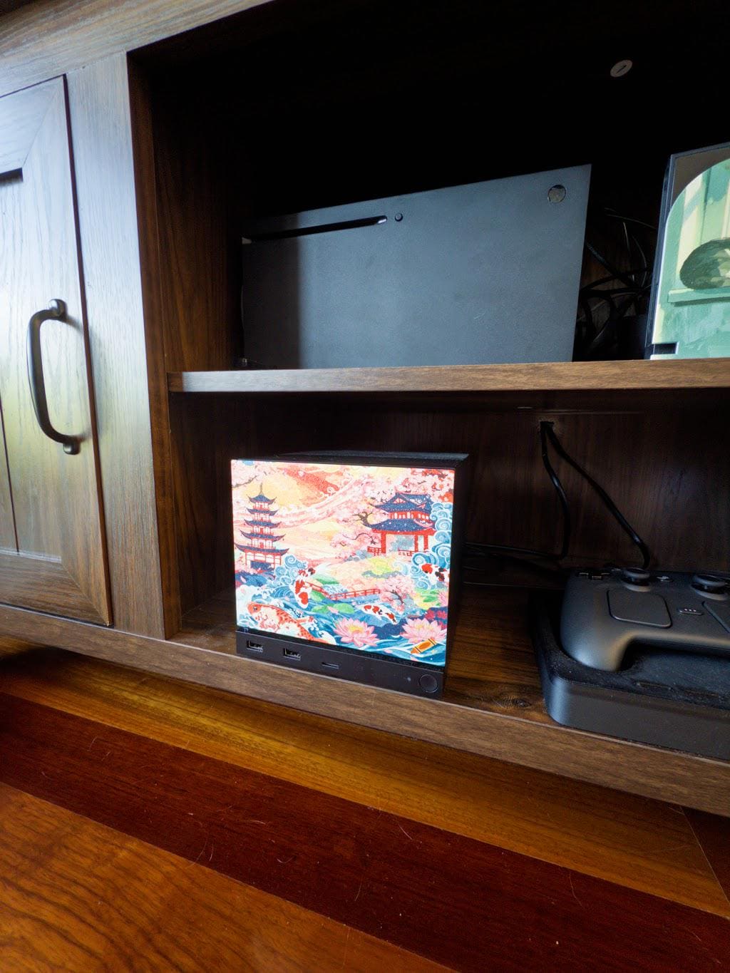 Koi Pagoda Garden Art | Steam Machine Faceplate | Cherry Blossom Sunset Panel | Magnetic UV Printed Cover | Japanese Gamer Gift with the cherry blossom pagoda art, angled side view installed on the console in a wooden shelf cubby beside a game controller